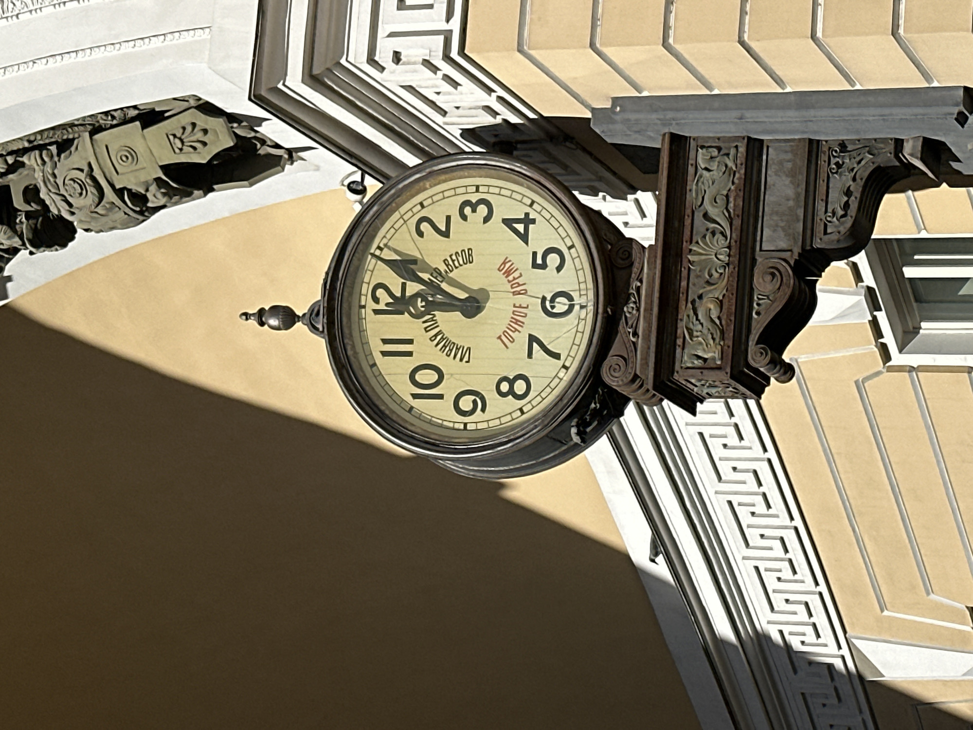 An eye-level shot captures a large, ornate clock mounted on the exterior corner of a grand building in Saint Petersburg, Russia. The image is rotated, showing the clock from a slightly upward angle, with architectural details framing it.
The circular clock face is light yellow with black Arabic numerals. Its hands point to approximately 11:55. Visible Cyrillic text on the clock face reads "ГЛАВНАЯ ПАЛАТА ВЕСОВ" (Main Chamber of Weights) and "ТОЧНОЕ ВРЕМЯ" (Exact Time), suggesting the clock's historical function related to standardized measurements. The clock is affixed to the building by a heavily decorated, dark metal bracket featuring intricate scrollwork and floral motifs.
The building's architecture is classical, characterized by creamy yellow walls and white decorative elements. To the left of the clock, partially obscured by shadow, is a sculpted stone figure, possibly an atlas or caryatid, with detailed facial features and headdress. To the right and above the clock, elaborate white architectural friezes with Greek key patterns and other geometric designs adorn the building's facade.
Strong, diagonal shadows cut across the building's warm yellow surface and the bottom right corner of the frame, indicating bright, sunny weather and likely mid-morning or early afternoon sun. No people or activities are explicitly depicted; the scene focuses on the static architectural details and the clock as a central object.