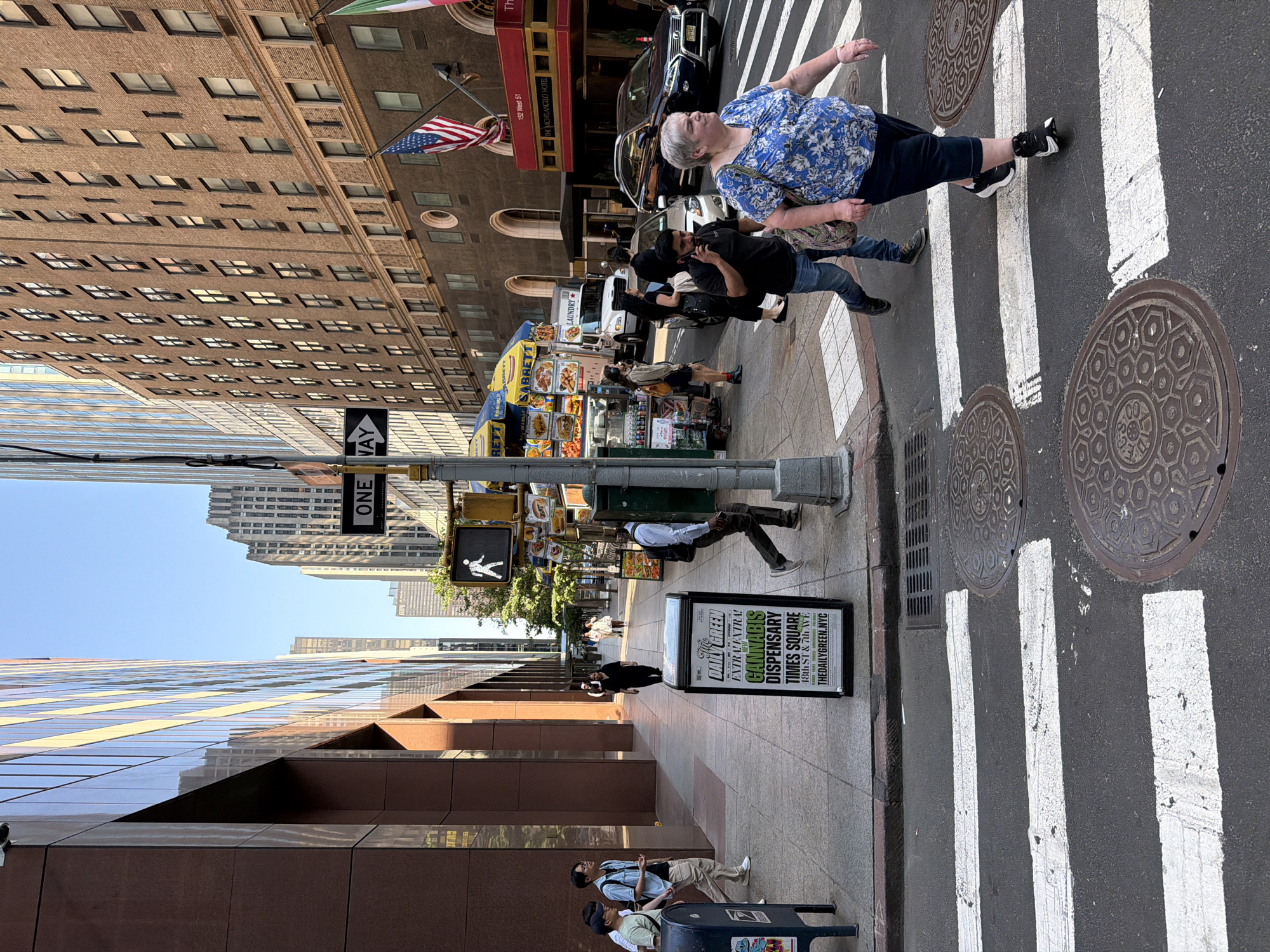 This photograph captures a street scene in New York City, United States of America. The view is from a slightly low angle, presenting a perspective of the city's buildings and street life.
The foreground features a pedestrian crosswalk marked with white stripes, adjacent to which is a sidewalk. A sidewalk advertisement for "The Daily Green Cannabis Dispensary" is seen. A trash can is visible on the right edge of the image, with a couple of people nearby.
On the crosswalk, an older woman is walking across the street. Near her, a man is walking and using a cell phone. Behind them, a hot dog food cart vendor is seen on the sidewalk.
The background is filled with the urban landscape of New York City. There are buildings of different architectural styles, with various windows and facades. Street signs are seen near the sidewalk. Also seen is the Michelangelo Hotel.