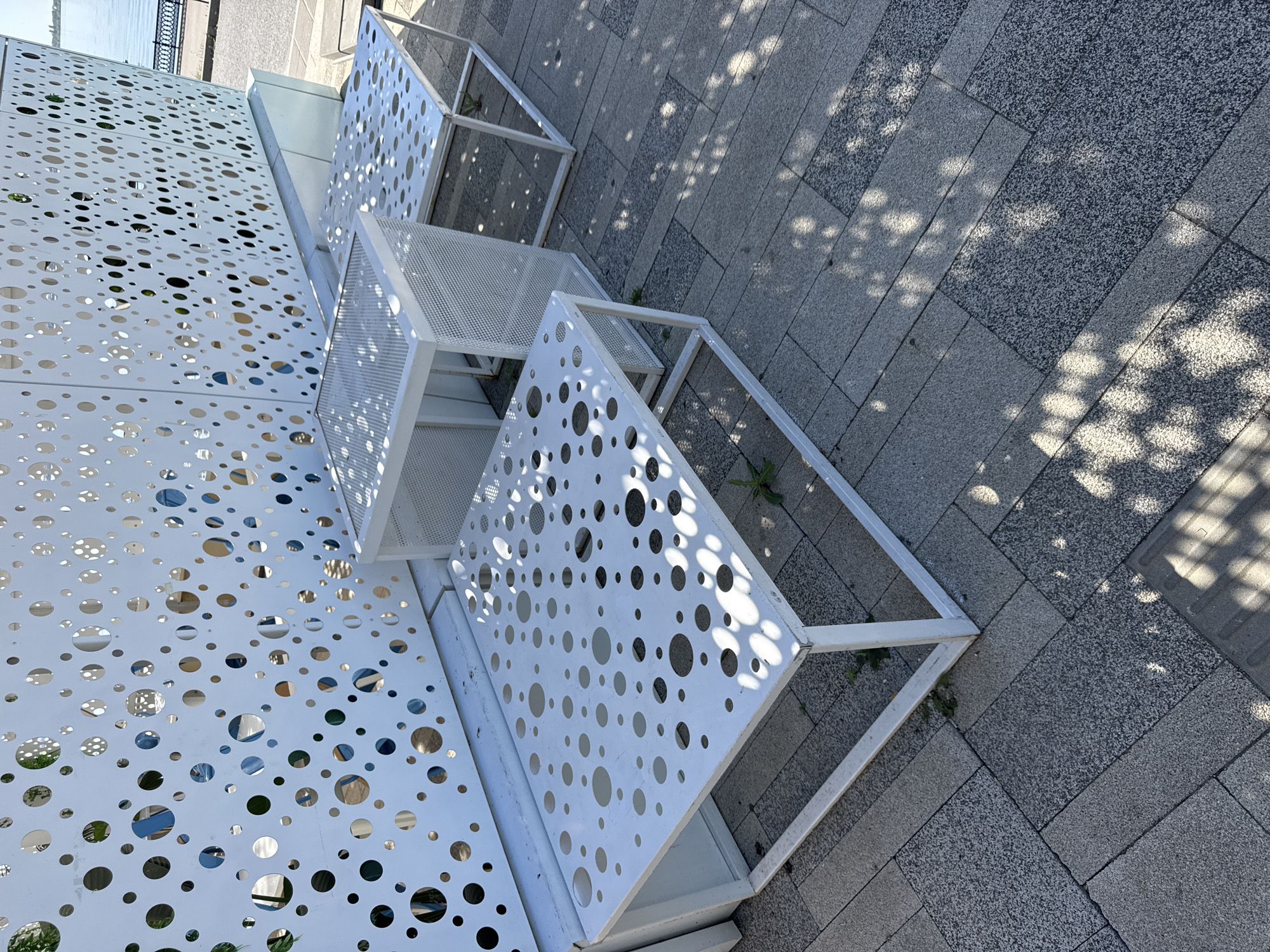 The image shows a section of the Saratov city embankment in Russia. There are white, cuboid metal structures arranged in a tiered manner, possibly forming part of street furniture or an art installation. The sides of these structures are perforated with round holes, and one of the middle tiers appears to have a fine mesh panel. The ground is paved with rectangular grey stones of varying textures. Tree shadows fall across the pavement. In the background, a body of water is visible, with a dark railing along its edge.
