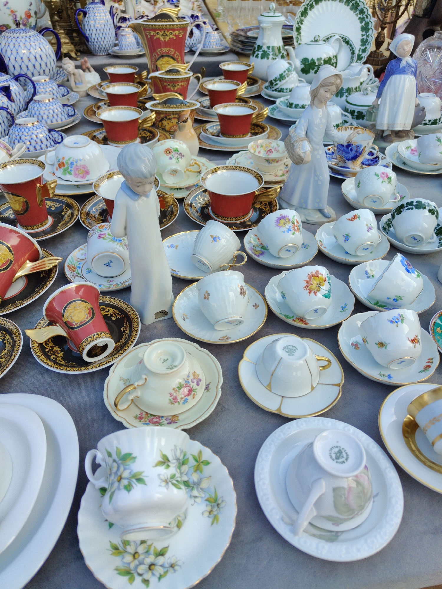 The image shows a table laden with a diverse collection of antique porcelain tableware, primarily teacups and saucers, as well as a few teapots and figurines. The items are arranged in a cluttered yet appealing manner, suggesting an outdoor market or antique stall.
The subjects are primarily inanimate objects: the porcelain cups and saucers display various intricate floral patterns, some with gold trim. There are also a few ceramic figurines of people, one a girl holding a basket and another a woman in traditional dress. The scene is set outdoors, likely in a marketplace in Gdańsk, Poland, given the location context. The lighting suggests it could be daytime, possibly late morning or early afternoon, under natural light. There are no explicit weather cues, but the general atmosphere feels like a sunny day at a market.
The activity taking place is the display and likely sale of these antique porcelain items. The arrangement of the items suggests they are being presented to potential buyers. There is no visible text in the image, other than what might be small maker's marks on the porcelain itself, which are too indistinct to read. The overall impression is one of abundance and the charm of collecting vintage decorative items.