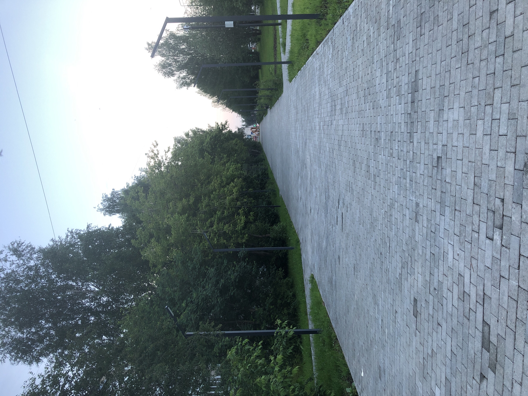 The image depicts a paved walkway lined with trees on either side, extending into the distance. Several modern black lampposts are positioned along the walkway. The sky is light blue, suggesting it is either early morning or late afternoon. In the far distance, a few figures can be seen, but they are too small to discern any details. The walkway is constructed from gray rectangular pavers. The overall scene appears to be a park or a well-maintained public area. The question implies a specific location in Solikamsk, Russia, however, no identifying markers are visible in the image to confirm this.