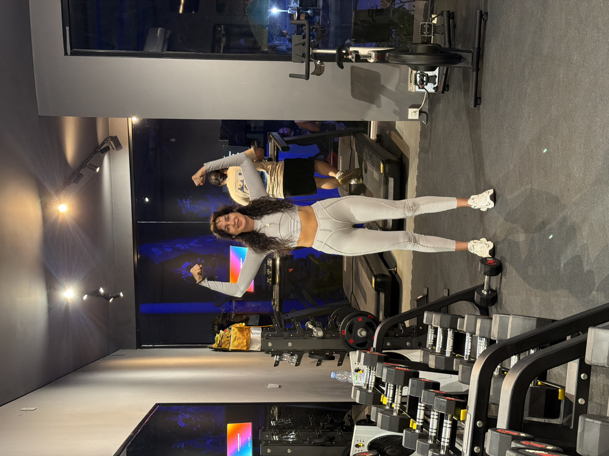 A woman with long, dark, curly hair is flexing her biceps in a gym. She is wearing a light gray athletic outfit, including a cropped long-sleeved top and matching leggings. Her arms are raised, and she is smiling at the camera. Behind her, a man with short, dark hair is jogging on a treadmill. The gym appears modern with dark gray walls and a dark floor. Equipment such as treadmills, weight racks with dumbbells, and a stationary bicycle are visible. The lighting is dim with spotlights on the ceiling and some colored lights on screens in the background. Through a large window on the right, an outdoor area with some seating and greenery can be seen, suggesting the gym might be in a resort or hotel.