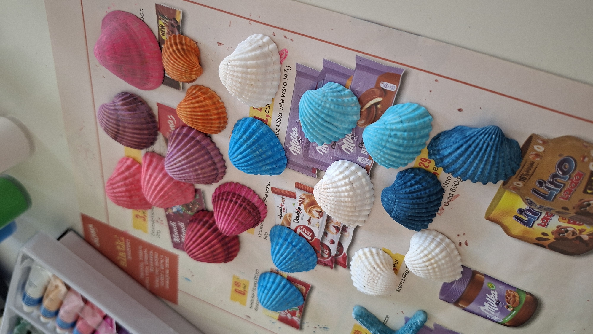 The image depicts a tabletop scene, likely an indoor setting, with various craft items and food packaging. In the upper left corner, a white box holds several tubes of paint in pastel colors, suggesting a crafting activity. Scattered across the surface are numerous seashells, painted in vibrant hues of pink, purple, orange, and blue, as well as natural white ones. Some of these seashells are arranged in clusters, while others are individually placed.
Beneath the seashells, several pieces of printed material are visible, including what appear to be product advertisements or flyers. These contain text and images of food products such as chocolate bars and jars of spread, with prominent branding like "Milka," "Hazelnut," and "Lino." The overall presentation suggests a crafting project involving decorated seashells, possibly for decoration or as part of an art project. The environment is brightly lit, with the surface appearing to be a flat, textured material, possibly a table or a large sheet of paper.
There is no discernible information within the image that would allow for the determination of its location within the city of Podgorica, Montenegro, or any other specific geographical location. The image focuses solely on the craft supplies and food packaging.