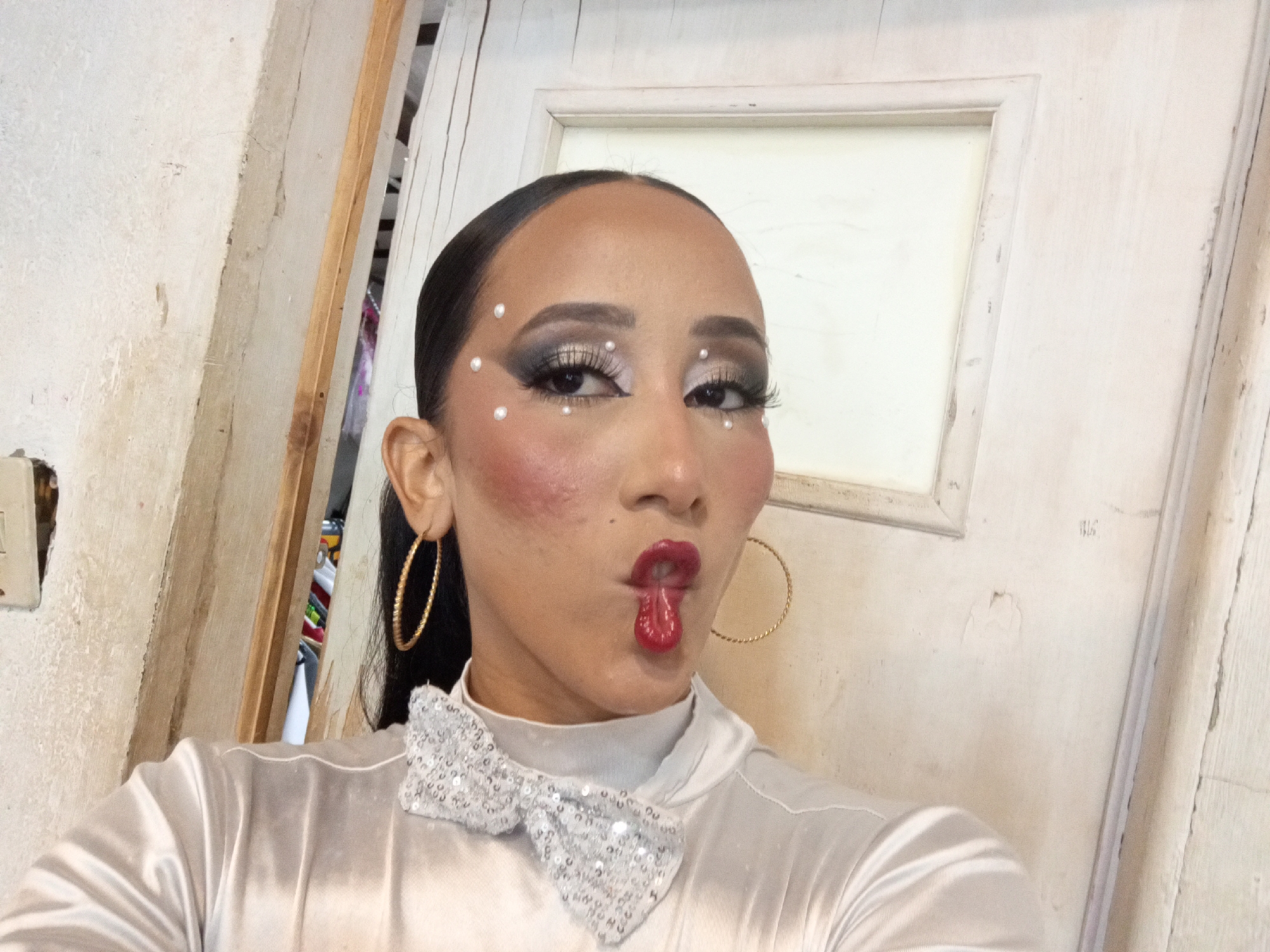 A person poses indoors against a backdrop of a white wooden door with a rectangular recessed panel. The person has dark hair slicked back, heavy eye makeup with faux eyelashes and small white dots as embellishments around the eyes and hair. The cheeks have heavy blush. Red lipstick is worn, and the person is making a fish-like mouth. The person wears large gold hoop earrings and a shiny silver bow tie on top of a white, high-necked satin top. The environment appears to be a residential interior with weathered walls and a wooden door. The location in the city of Porlamar, Venezuela cannot be determined from this image alone.
