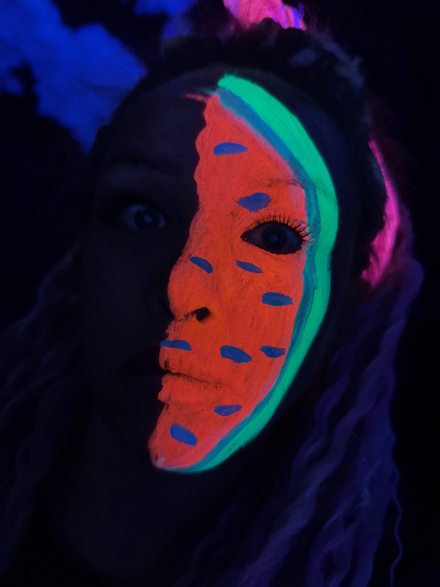 A person with neon face paint is lit with a black light in a dark environment. Half of the face is visible and painted to resemble a watermelon, with red paint, blue "seeds," and a green rind. The other half of the face is shadowed. The person has long hair in two colors, and appears to be surprised. The city of Noviy, Russia is not relevant to the image.