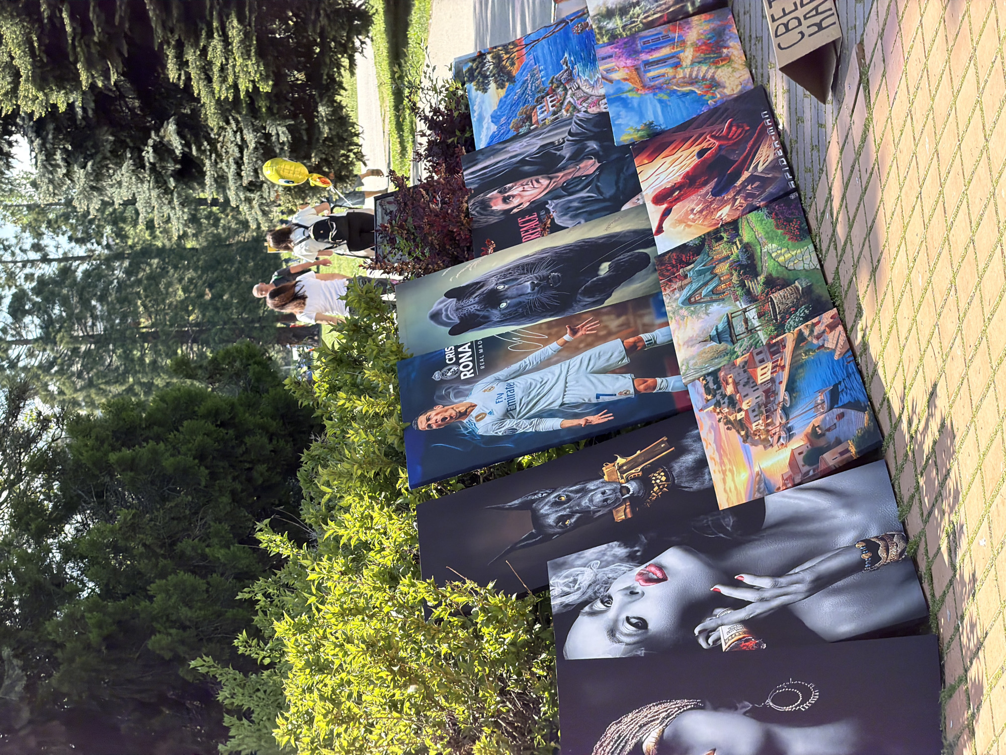 Here is a forensic, matter-of-fact description of the image:
The image shows a collection of prints or paintings displayed on a brick-paved surface, possibly for sale. The subjects of the artwork vary and include portraits of people, animals, landscapes, and fictional characters (e.g., Spider-Man). There is a print of Cristiano Ronaldo in a Real Madrid jersey.
In the background, there are people walking on a path, partially obscured by trees and foliage. A yellow balloon is visible among the people.
The location is likely an outdoor area with trees and paved walkways, which is consistent with an outdoor art market or display area in Sofia, Bulgaria. A piece of cardboard with Cyrillic script is visible near the artworks.