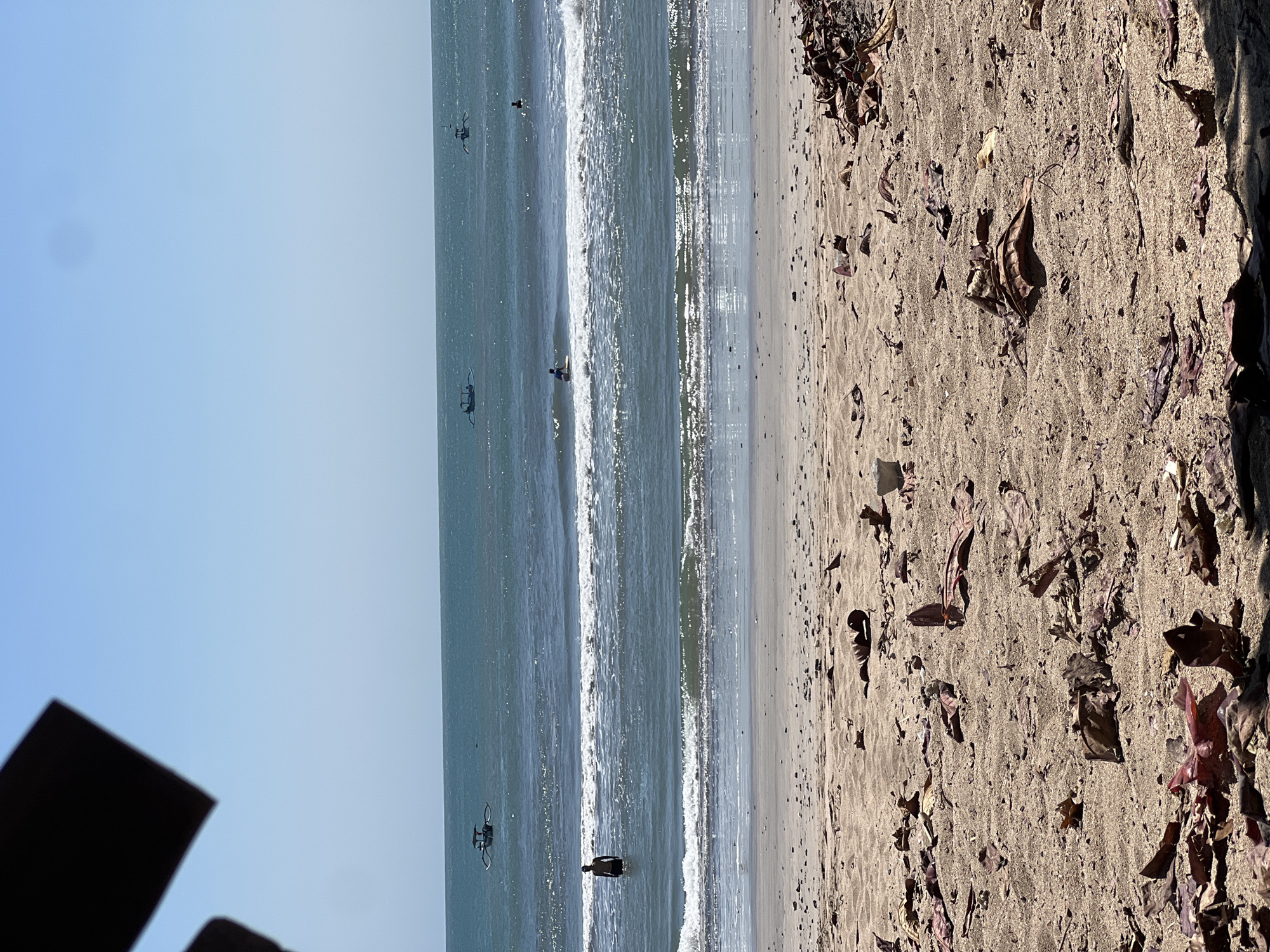 A bright, sunny daytime shot captures a serene beach scene in Kuta, Indonesia. The foreground is dominated by a sandy beach scattered with dried leaves and some debris, suggesting a natural, unmanicured environment. Beyond the beach, the ocean stretches out, with gentle waves breaking near the shore. Several people are visible in the water, some wading and one appearing to surf. A few traditional outrigger boats are dotted across the expanse of the sea, indicating local marine activity. The sky is a clear, pale blue, creating a tranquil backdrop for the coastal landscape. The overall impression is one of a peaceful day at the beach.