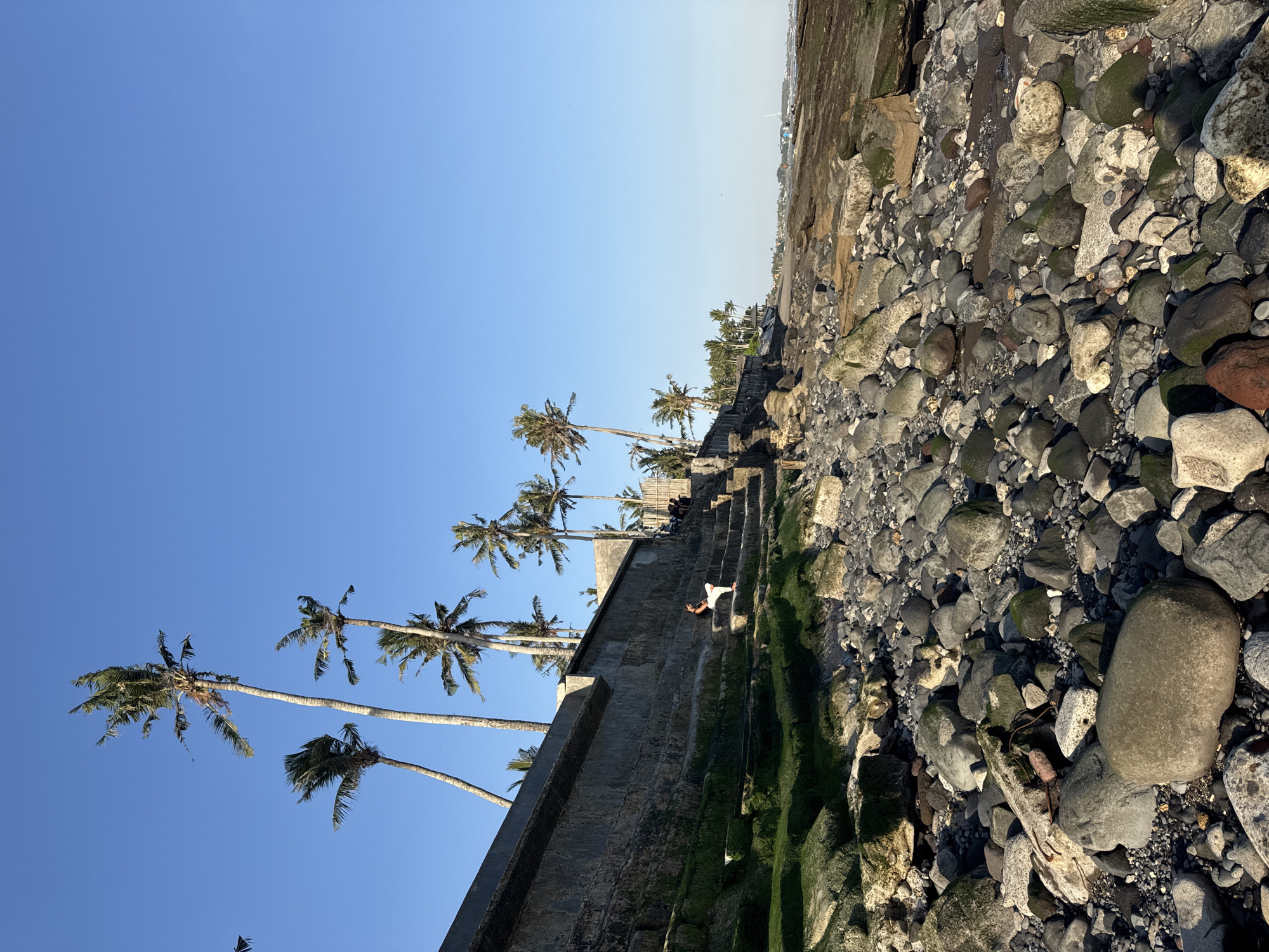 A long, low-angle shot captures a rocky beach on the coast of Mengwi, Indonesia, under a clear, bright blue sky. In the foreground, a jumble of smooth, grey, and dark rocks of various sizes fill the lower half of the frame, with some mossy green patches visible on larger stones. A concrete wall, made of rough-hewn stone blocks, runs along the left side of the image, forming a raised embankment. Steps, also made of stone and covered in green moss, ascend the embankment. A person is seated on one of these steps, facing to the left, with their legs crossed. Behind the wall, several tall palm trees with sparse fronds reach towards the sky. The background shows more palm trees, a distant shoreline with a few buildings, and the ocean stretching to the horizon. The lighting suggests it is either early morning or late afternoon, casting long shadows.