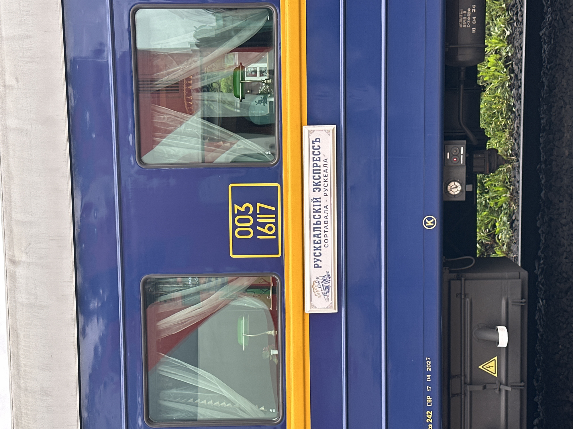 The image shows the side of a blue train car with a yellow stripe. There are two windows, each with red and white curtains. A sign on the train reads "РУСКЕАЛЬСКИЙ ЭКСПРЕССЬ" (Ruskeala Express) with "СОРТАВАЛА - РУСКЕАЛА" (Sortavala - Ruskeala) below it. Below the yellow stripe, there is a black box with a yellow triangle and lightning bolt symbol. Further to the right, there is a circular gauge and some technical markings on the train car. The train is on tracks, with gravel and green grass visible on the sides. The overall impression is of a well-maintained train car, likely part of a tourist route between Sortavala and Ruskeala.
