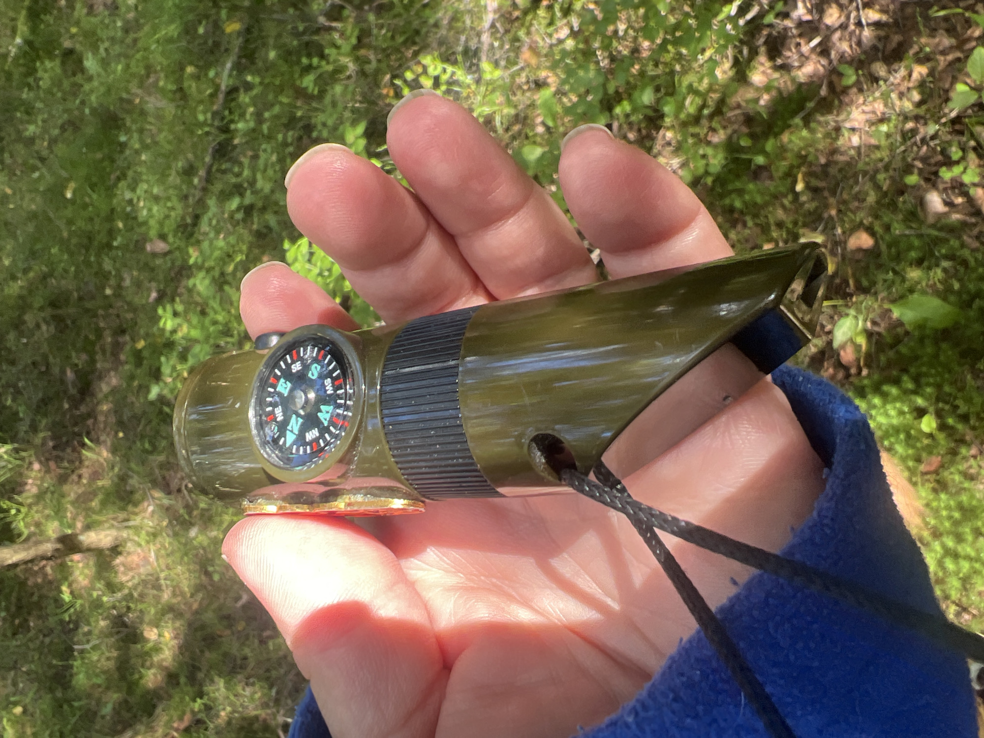 A hand holds a green, cylindrical object with a compass on one end. The compass has a black and white dial with directional markings. The object appears to be made of plastic and has a ridged section. A black cord is attached to the object. The background is blurred and shows green foliage, suggesting an outdoor environment. The lighting indicates it is daytime.