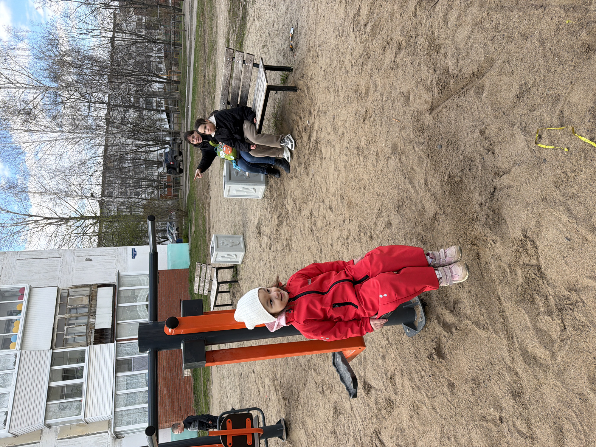 The image depicts an outdoor scene at a playground in Chaikovsky, Russia. The ground is covered in sand. A young girl in a red snowsuit and a white beanie is lying on an orange and black exercise machine. Two women are sitting on a bench in the background, with one pointing towards something. The weather appears to be overcast. A multi-story residential building is visible in the background.