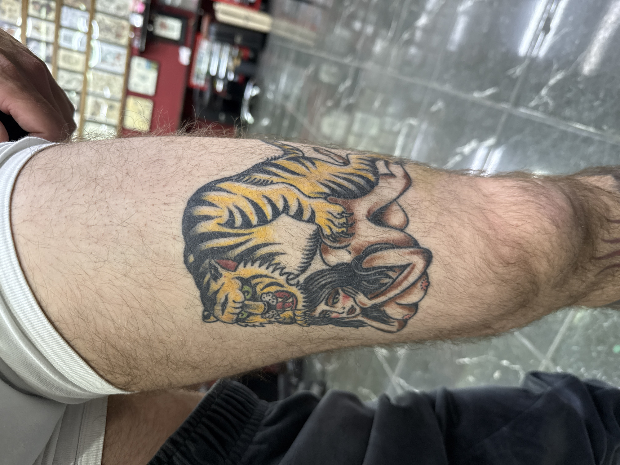 A close-up, low-angle shot focuses on a tattoo of a tiger appearing to be on top of a nude woman, depicted in a traditional tattoo style. The tattoo is on the hairy skin of a person's thigh, with the top of white underwear visible at the upper left and dark fabric, possibly athletic shorts, visible on the left. The background is out of focus but appears to be the interior of a tattoo parlor, with walls lined with framed artwork and display cases. The floor is shiny and reflective. The lighting is somewhat dim, suggesting an indoor setting. The image does not provide any specific information about the city of Moscow, Russia.