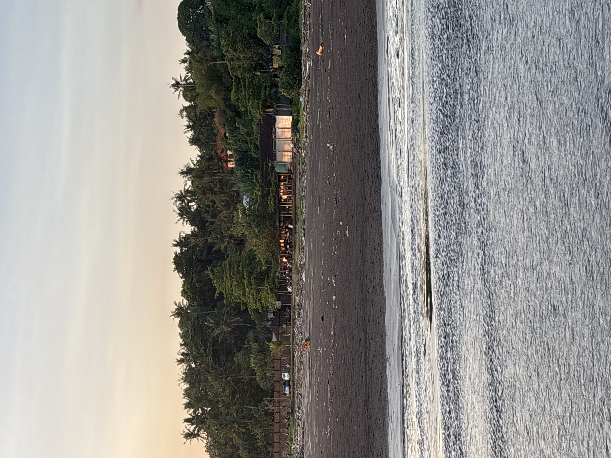 The image depicts a coastline, likely a beach, in Kerambitan, Indonesia. Lush green vegetation, including palm trees, lines the edge of the beach, with structures, possibly restaurants or resorts, visible among the trees. The beach itself is dark sand, meeting the water's edge where waves break. In the distance, the sky is a pale, muted color, suggesting either sunrise or sunset. There are a few cars are seen parked along a side road. The lighting indicates it could be early morning or late afternoon.