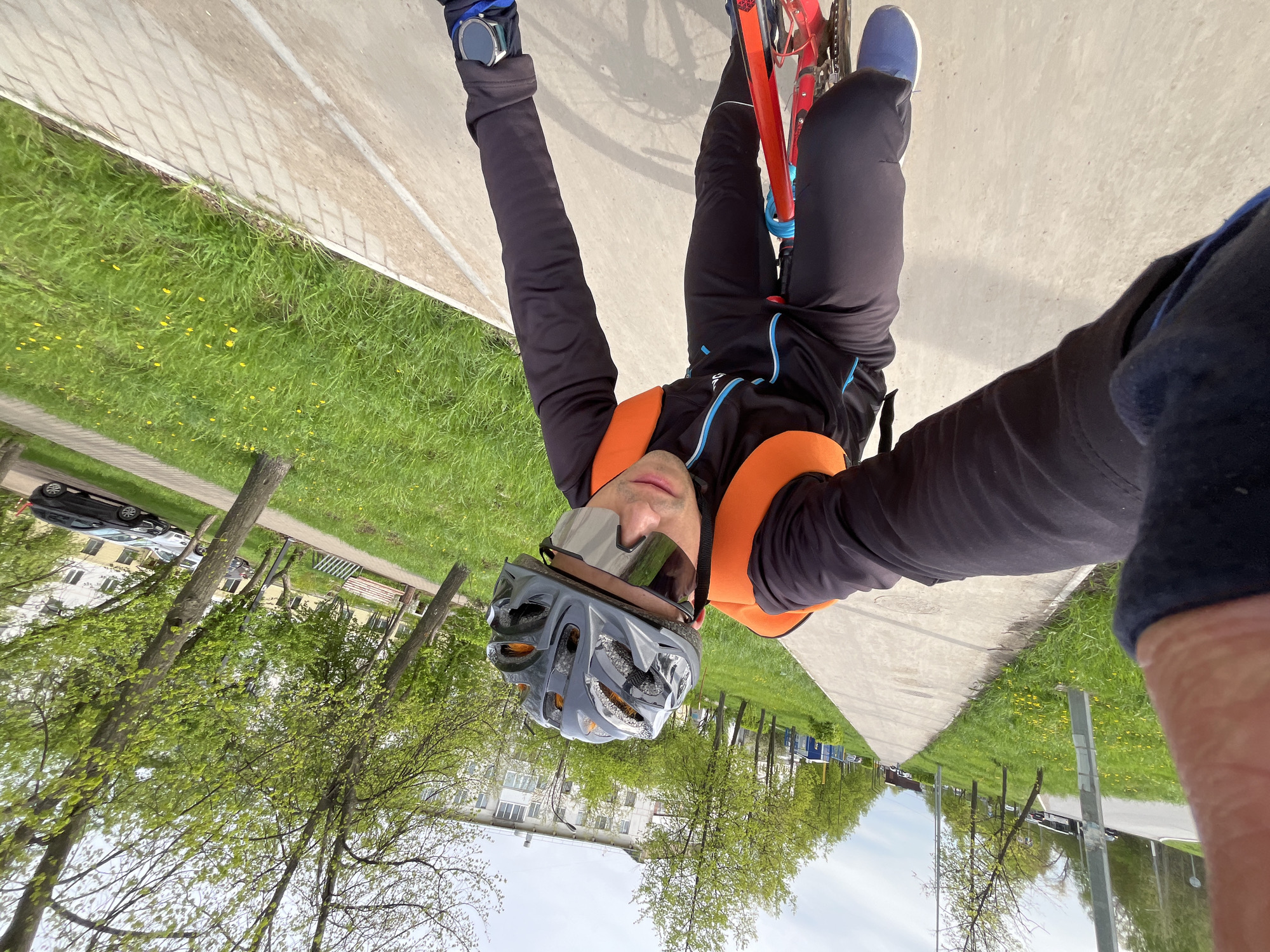 The image is an upside-down selfie taken by a man riding a bicycle on what appears to be a sidewalk next to a grassy area. The man is wearing a gray helmet, sunglasses, a black and orange athletic jacket, black pants, blue shoes, and a wristwatch. He is holding the handlebars of the bicycle, which has a red frame. In the background, there are trees, a building, and parked cars. The location appears to be in Chaikovsky, Russia, based on the urban environment and vegetation.