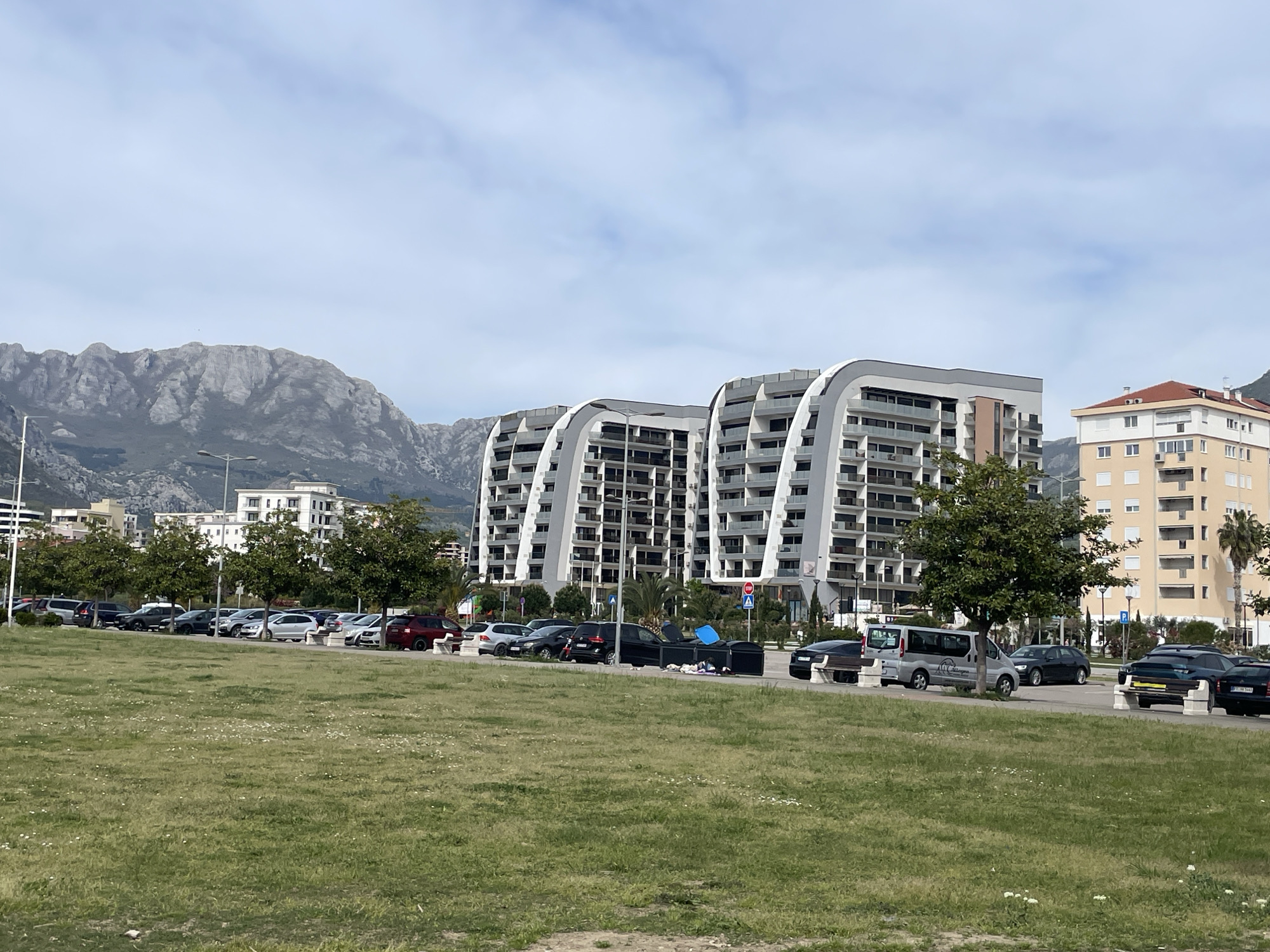 The image captures an urban landscape in Bar, Montenegro, featuring modern apartment buildings with curved architectural elements against a backdrop of mountainous terrain. A grassy field occupies the foreground, leading to a row of parked cars and trees that line the street. The scene includes a mix of residential and commercial buildings, suggesting a populated area. The sky is partially covered with clouds.