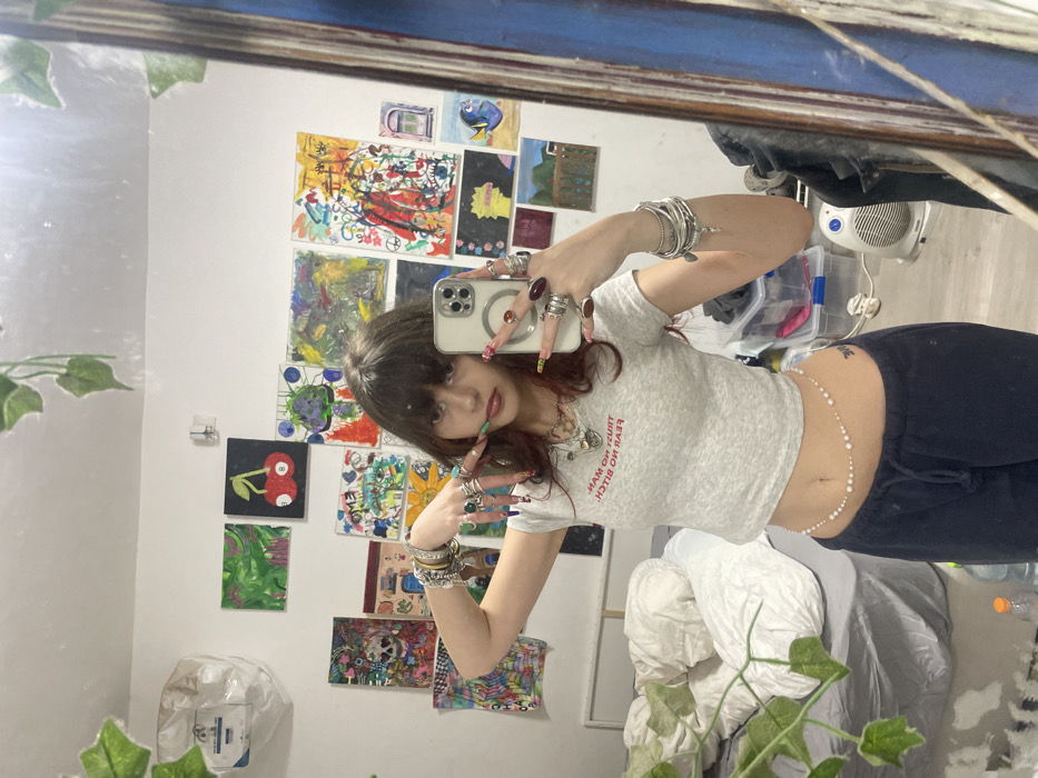 A young person with dark hair and bangs is captured in a mirror selfie within a bedroom in Raanana, Israel. The individual wears a light grey fitted crop top with red text reading "TRUST NO MAN, FEAR NO BITCH," dark blue drawstring sweatpants, and a white beaded waist chain. They are heavily accessorized with numerous silver-toned rings on both hands, multiple silver-toned bracelets on the right wrist, and several necklaces. The fingernails are long, artificially extended, and decorated with diverse colors and patterns. A small dark-ink tattoo is visible on the lower left abdomen. The person holds a light-colored smartphone, equipped with multiple camera lenses, in their left hand, reflecting the action of taking the photo.
The background, visible through the mirror's reflection, consists of a white wall largely covered with various pieces of artwork, including abstract paintings, a skull motif, a cherry billiard ball, character depictions resembling Lisa Simpson and Dory, and a landscape. Artificial green foliage is draped near the top edge of the mirror and potentially along the wall. To the left, a bed with crumpled white bedding is present. On the floor to the right, a clear plastic storage bin, a white electric fan, and a clear plastic bottle containing an orange liquid are visible, along with assorted cables. A dark-colored, possibly wooden, mirror frame runs along the right edge of the image.