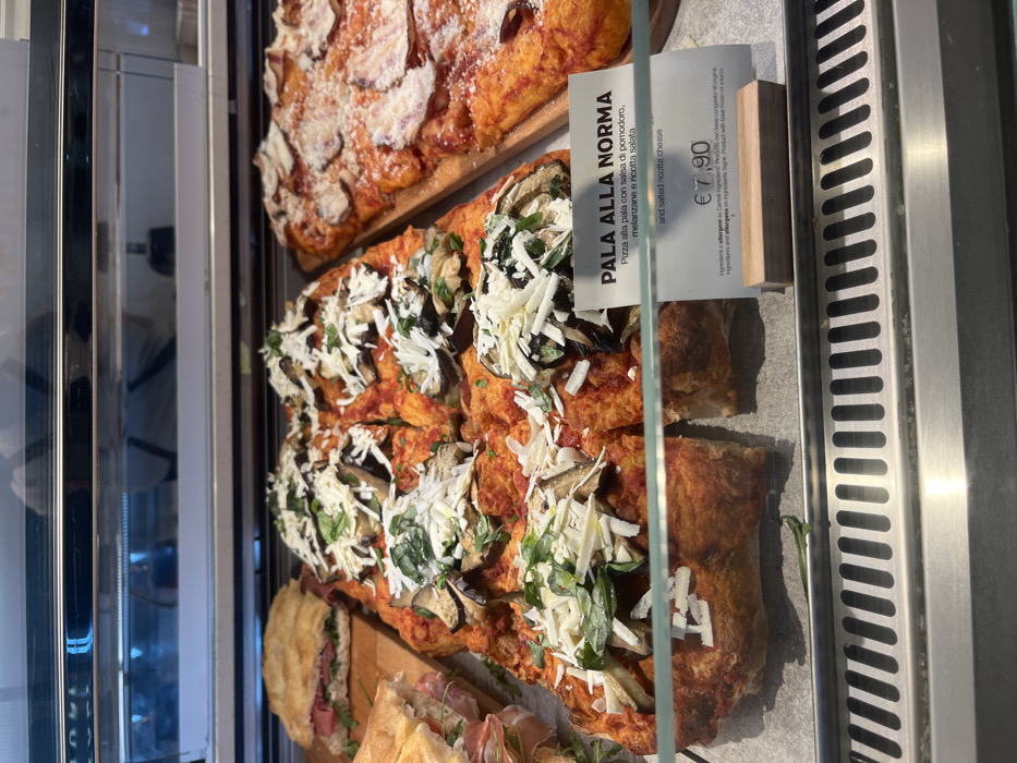 The image displays a vibrant selection of food items, likely in a deli or bakery counter in Dorno, Italy. The primary focus is on two rectangular flatbread pizzas, known as "Pala alla Norma," arranged in a refrigerated glass display case.
The pizza prominently featured in the foreground is generously topped with a rich tomato sauce, slices of eggplant, fresh green herbs (likely basil), and a generous scattering of shredded white cheese, which the label identifies as salted ricotta. This pizza has been cut into several individual portions, indicating it's ready for sale by the slice. Above it, another rectangular pizza is visible, adorned with what appears to be thin slices of cured meat or cheese, dusted with white powder, possibly grated hard cheese. Below these main items, other food products, including sandwiches or further flatbreads, can be partially seen.
A white price tag with black text is clearly visible, describing the foreground pizza. It reads: "PALA ALLA NORMA," followed by "Pizza alla pala con salsa di pomodoro, melanzane e ricotta salata" (Flatbread pizza with tomato sauce, eggplant, and salted ricotta). Below this, "and salted ricotta cheese" provides an English translation. The price listed is "€ 7,90." Further down, text in Italian and English states: "Ingredienti e allergeni su Cartelli Ingredienti. Prodotto con base congelata all'origine." and "Ingredients and allergens on Ingredients Signs. Product with base from frozen at source."
The scene suggests a casual eatery or food market, with the food presented appealingly for customers. The metallic elements of the display case, including a grated vent system on the right, add to the commercial setting. The lighting is bright, illuminating the food clearly.