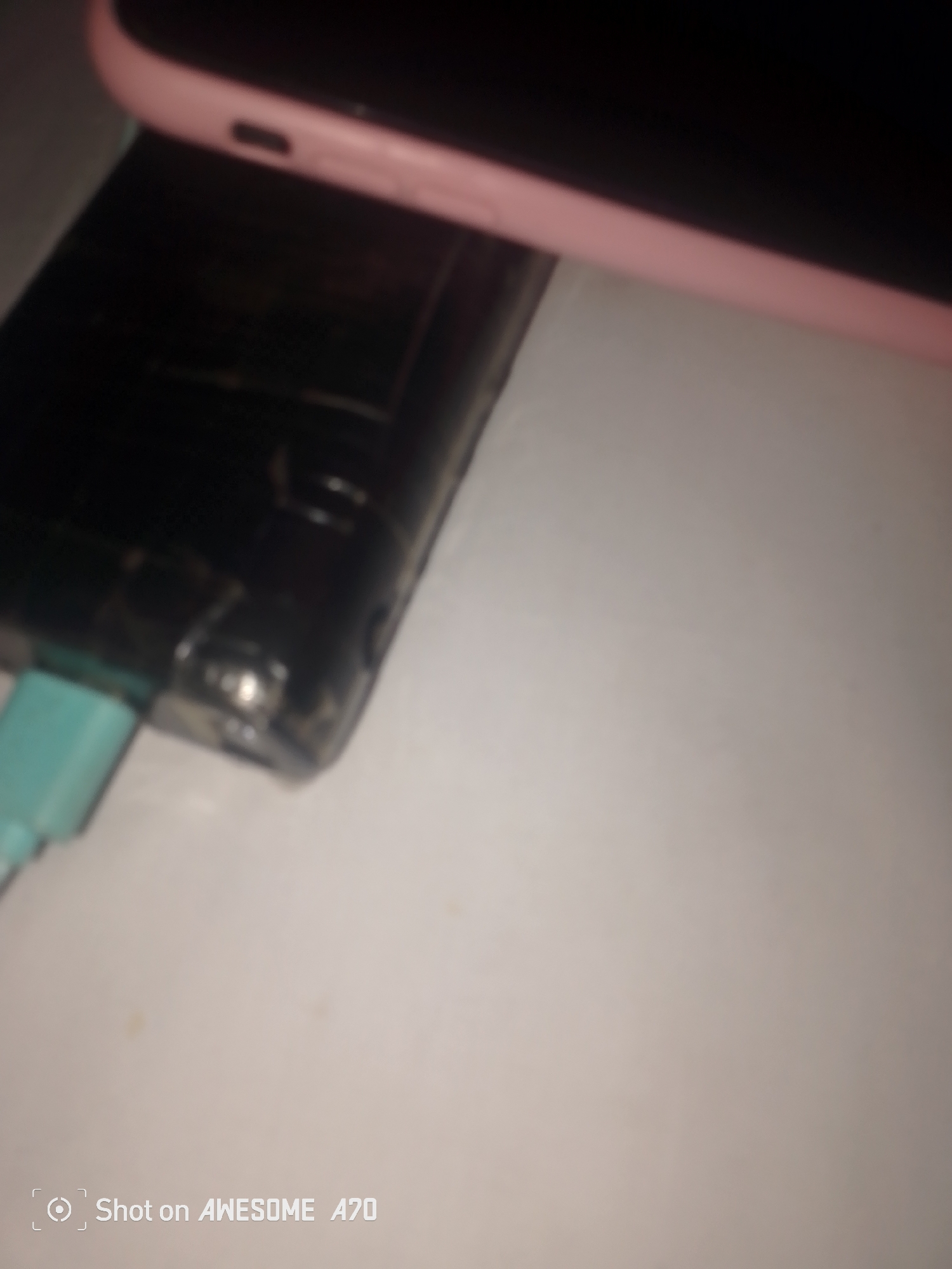 The image shows a close-up, slightly blurred view of a pink-cased electronic device resting on a black rectangular object, likely a power bank or external battery. A light blue charging cable is plugged into the black object. The scene is set on a light-colored surface, possibly a table. A watermark in the bottom left corner reads "Shot on AWESOME A70." There is no indication of Jauro Yinu, Nigeria in the image.