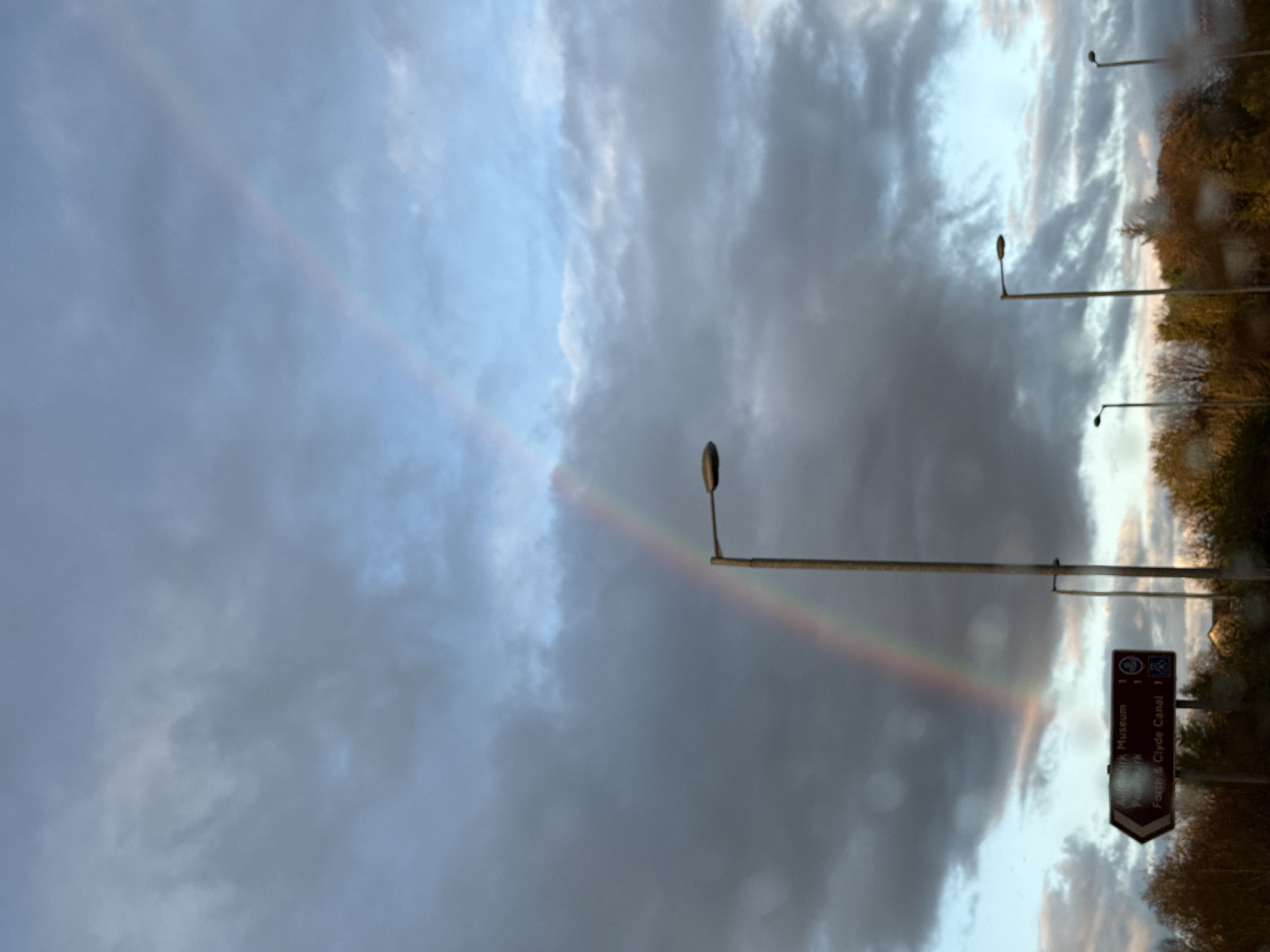 The image captures a sky filled with gray and white clouds, with a faint rainbow arc visible. Streetlights are present, extending into the frame from the right side, with a road sign partially visible. The sign indicates "Park Museum 1" and "Forth & Clyde Canal 1". The location is in the city of Unknown, Unknown.