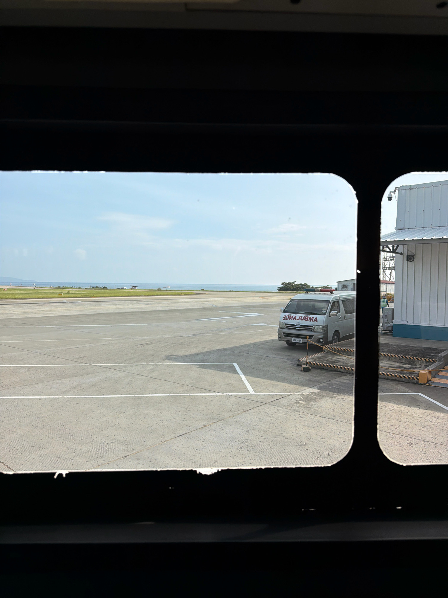 The image shows an exterior view of an airport tarmac through a dark-framed window. In the foreground, the dark frame of the window is visible, with the bottom edge appearing rough and damaged. The middle ground features a vast, grey tarmac with white markings indicating lanes or taxiways. In the background, a body of water, likely the sea, is visible under a pale blue sky with scattered white clouds.
On the right side of the tarmac, a white ambulance vehicle with flashing red lights on its roof is parked. The word "AMBULANCE" is clearly visible on the side of the vehicle, written in reverse. To the right of the ambulance, part of a white building with a corrugated roof and blue trim is visible. A security camera is mounted on the building. Yellow and black striped barriers are placed on the ground in front of the building. A person in a light-colored uniform and a yellow vest is standing near the building. Further in the distance, a few small structures and some greenery can be seen near the water's edge. The overall scene suggests an active airport environment. The location is identified as the vicinity of Caticlan Airport (MPH) in Malay, Aklan, Philippines, which serves the island of Boracay and the surrounding areas.