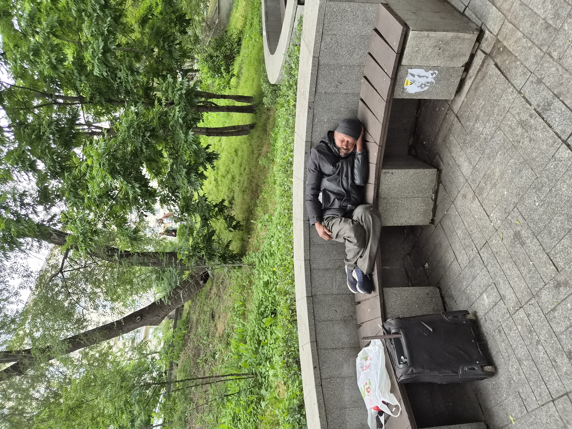 A man sleeps on a park bench in Vladivostok, Russia, with his belongings beside him. The man is lying on his side, his head resting on his arm. He is wearing a black jacket, dark pants, and a black beanie. To his left, there is a black suitcase with wheels and a white plastic shopping bag. The bench is made of wood and sits on a stone base. Behind the bench is a grassy slope with trees and some buildings in the background. The ground in front of the bench is paved with gray tiles.