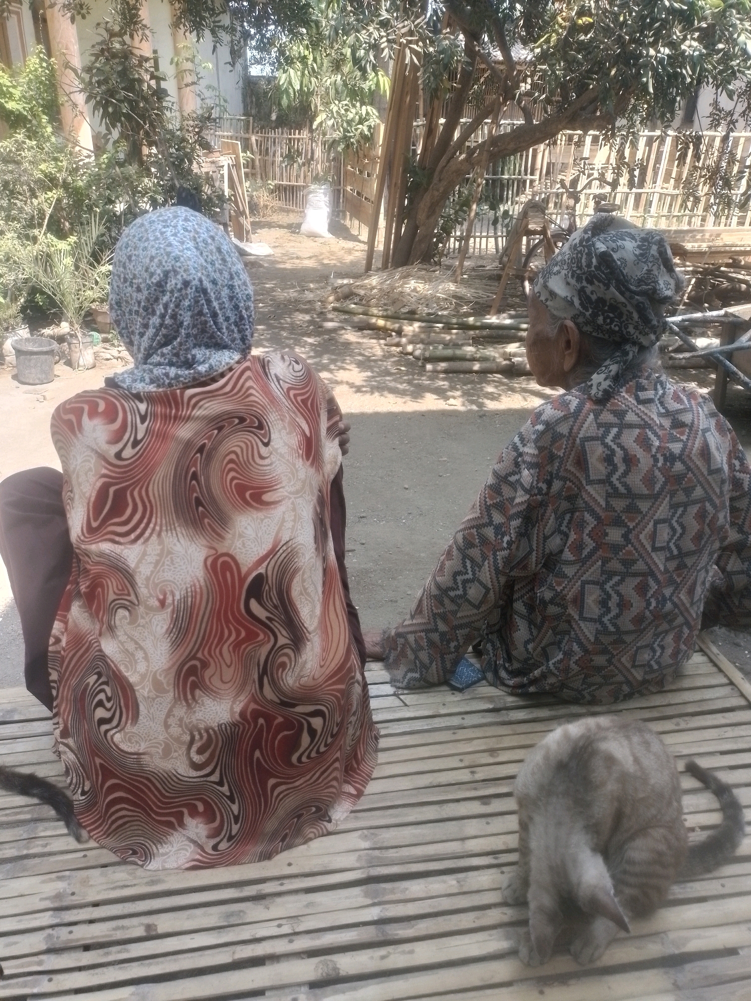 Two elderly individuals are seated on a bamboo mat outdoors. The person on the left, seen from the back, wears a patterned headscarf and a blouse with a swirling red and brown print. They are wearing brown trousers. The person on the right, also seen from the back, wears a patterned headscarf and a patterned blouse with geometric designs in brown, black, and tan. A grey cat is curled up on the bamboo mat in front of them. The background shows a yard with trees, a bamboo fence, and some construction materials. The lighting suggests it is daytime.