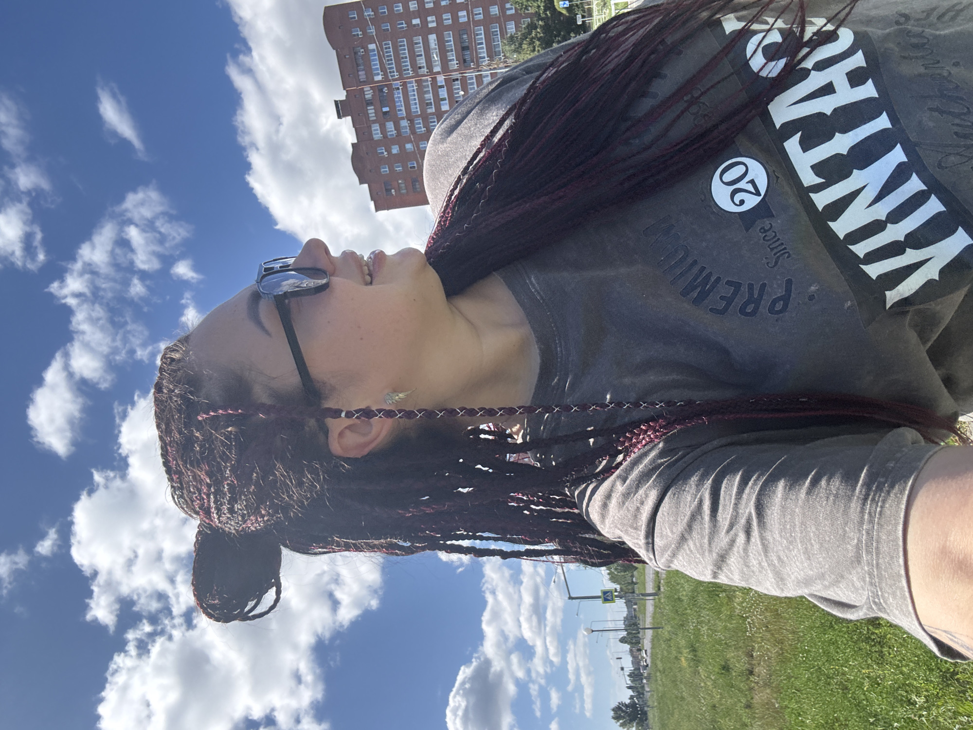 A woman with dark braided hair, some of which is adorned with burgundy threads, is captured from the chest up, looking to her right. She is wearing sunglasses and a gray t-shirt with the words "PREMIUM Since 20 VINTAGE" printed on it. She is outdoors on a sunny day with a blue sky filled with white clouds. In the background, a tall, reddish-brown brick apartment building is visible, along with a road and green grassy areas. The location is the city of Tolyatti, Russia.