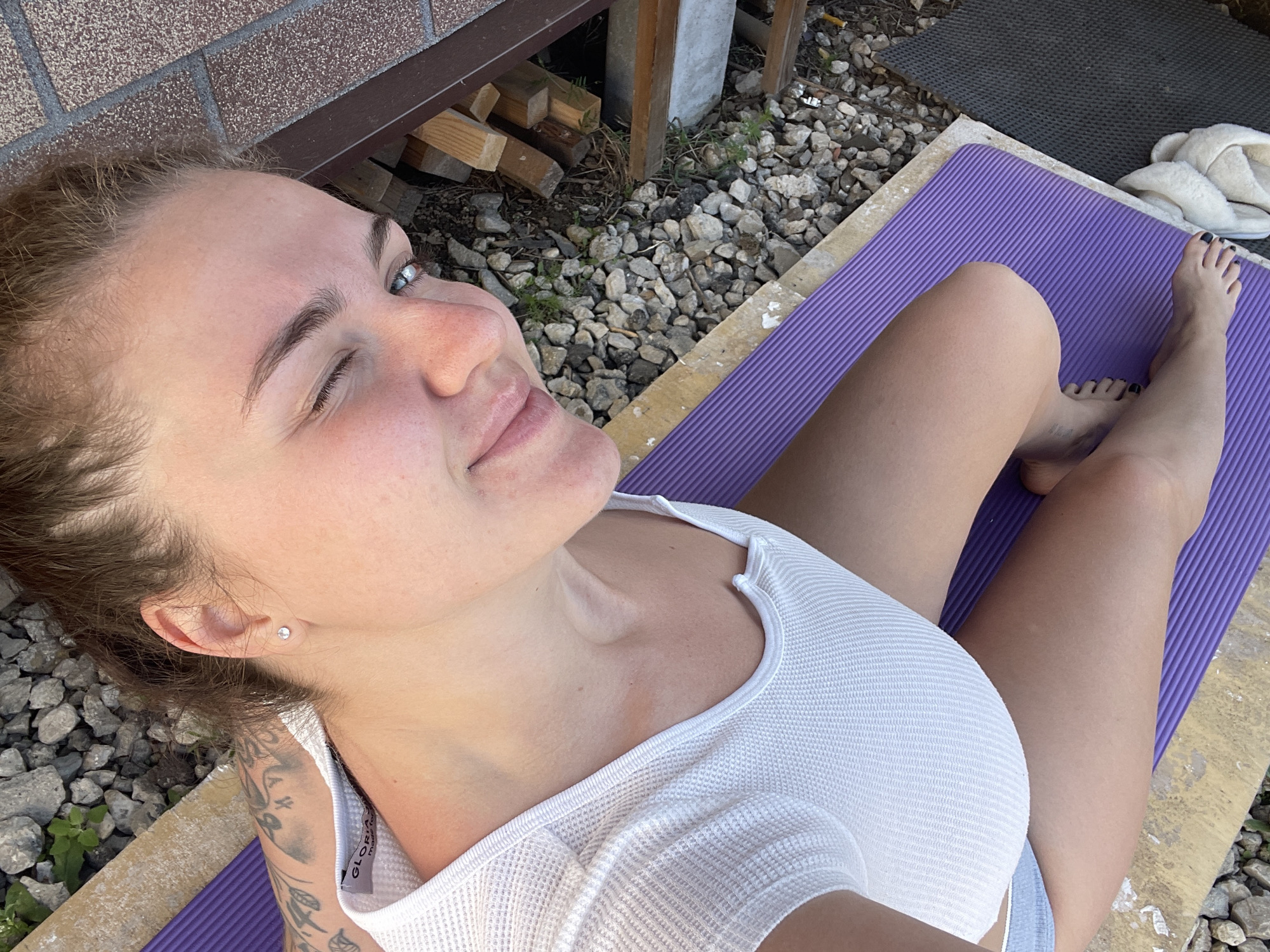 A woman is lying on a purple yoga mat outdoors. She is wearing a white tank top and has a tattoo on her left arm. Her toes are painted black. The location appears to be a backyard or patio area with gravel, a brick wall, and wooden planks. There is a gray mat and white slippers near her feet. The photo is taken from a high angle, looking down at the woman. The woman is located in the city of Мосметаллоконструкция Онт, Russia.