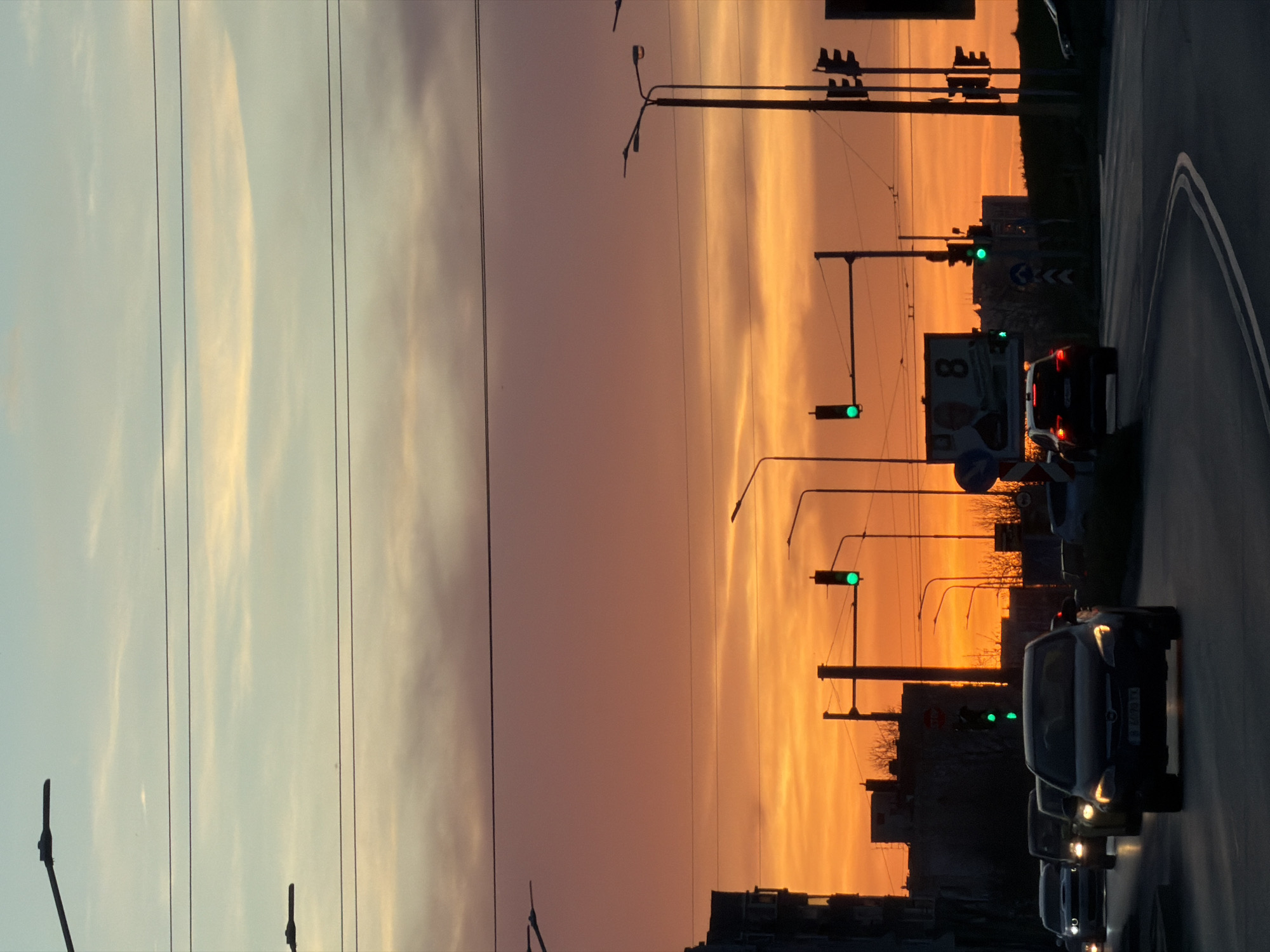 The image shows a sunset scene on a road in Varna, Bulgaria. The sky transitions from light blue to orange and red near the horizon. There are utility poles and power lines visible against the sky. The road has multiple lanes and shows several cars driving with their headlights on. There are traffic lights that are showing a green signal. Buildings and trees can be seen in silhouette against the horizon.