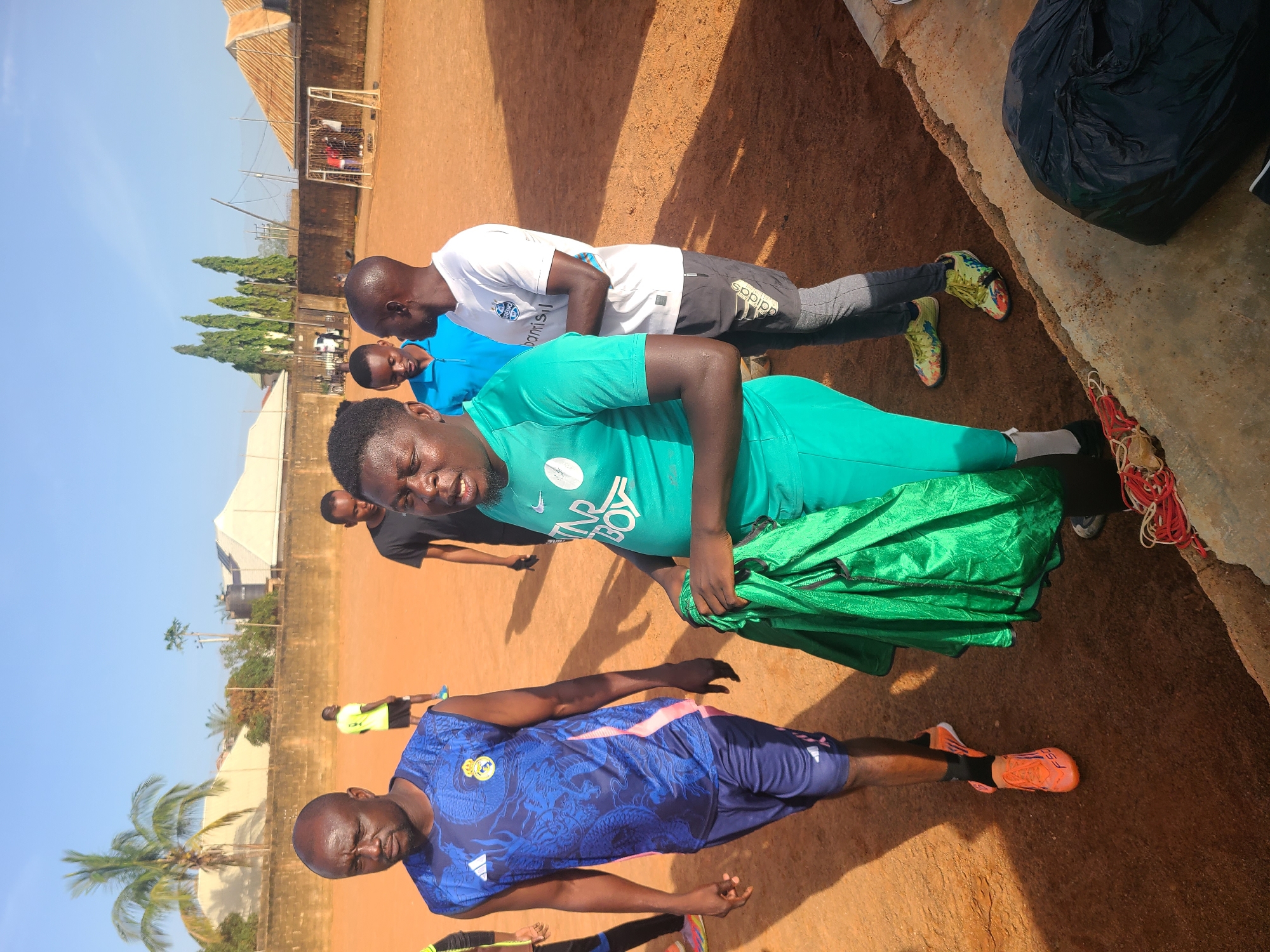 The image depicts an outdoor scene at what appears to be a football field in Barakin Lerawi, Nigeria. The ground is covered in reddish-brown dirt. In the foreground, a group of men are visible. One man on the left is wearing a blue sleeveless football jersey with a dragon design and navy blue shorts. To his right, another man is wearing a bright green football jersey and shorts, holding a folded green fabric. Behind him, a man in a blue t-shirt is partially visible, and further back, a man in a black t-shirt. On the right side of the image, a man in a white t-shirt and grey leggings with black shorts is standing. In the background, a brick wall runs along the perimeter of the field, with some buildings and trees beyond. A football goal is visible in the far right background, with a few people standing near it. The sky is clear and blue, suggesting a daytime setting.