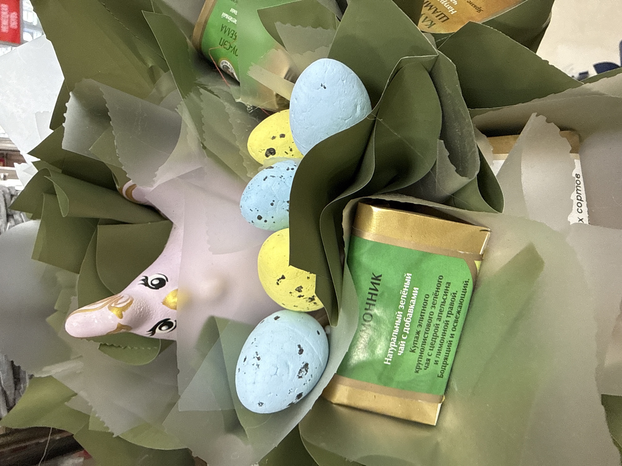 The image displays a close-up, high-angle view of a gift arrangement, situated within an indoor retail setting in Dmitrov, Russia. The central subject is a collection of items artfully presented within multiple layers of olive-green and semi-translucent wrapping paper, resembling a nest.
Dominating the upper portion of the arrangement is a light pink ceramic figure, stylized as a chick or small bird. It features prominent black and white eyes with painted eyelashes, a small yellow beak, and subtle gold detailing on its head. Five decorative, speckled, egg-shaped objects are nestled among the green wrapping. Three of these eggs are light blue with dark brown speckles, and two are yellow with dark brown speckles.
In the foreground, a rectangular package with a glossy gold wrapper and a distinct green label is visible. The Cyrillic text on this label translates to "MOLOCHNIK Natural green tea with additives," further specified as "a blend of elite large-leaf green tea with orange zest and lemongrass, invigorating and refreshing." To the right, a second, partially obscured package with a green and gold label is present, showing fragments of Cyrillic text including "Natur" and "Bright." Another partially visible label at the bottom includes the Cyrillic phrase "x sorts," indicating varieties.
The background is out of focus, suggesting an interior space with indistinct shapes consistent with retail shelving or displays. A blurred red sign with white Cyrillic text is visible in the upper right background. No individuals are depicted in the scene.