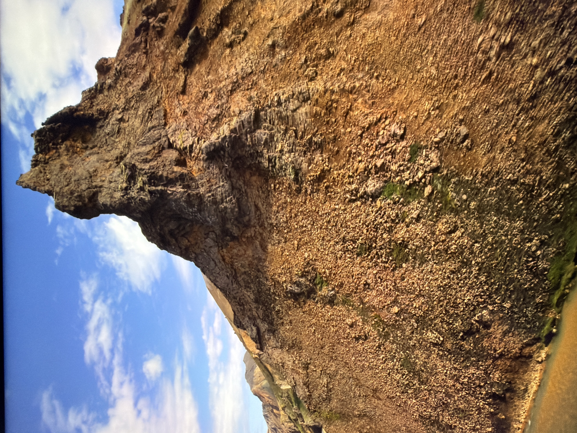 The image displays a steep, rocky mountainside under a bright blue sky with scattered white clouds. The terrain is predominantly covered in loose scree and larger rocks, with patches of green vegetation visible in some areas. A prominent, jagged rock formation rises sharply in the upper portion of the image. In the lower left corner, a body of water, likely a river or lake, with a yellowish-brown hue is visible. The overall scene suggests a natural, undeveloped landscape. There are no people, man-made structures, or any indication of urban environments present in this image. Therefore, this image cannot be associated with the city of Kyiv, Ukraine.