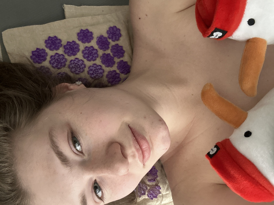 A fair-skinned woman reclines on a light brown mat adorned with violet floral-patterned circles. She faces the camera with a slight smile. Her eyes are light-colored, and she wears a small earring. Adjacent to her on the right are two white plush ducks with red caps and orange beaks. The scene appears to be indoors, illuminated by natural light.