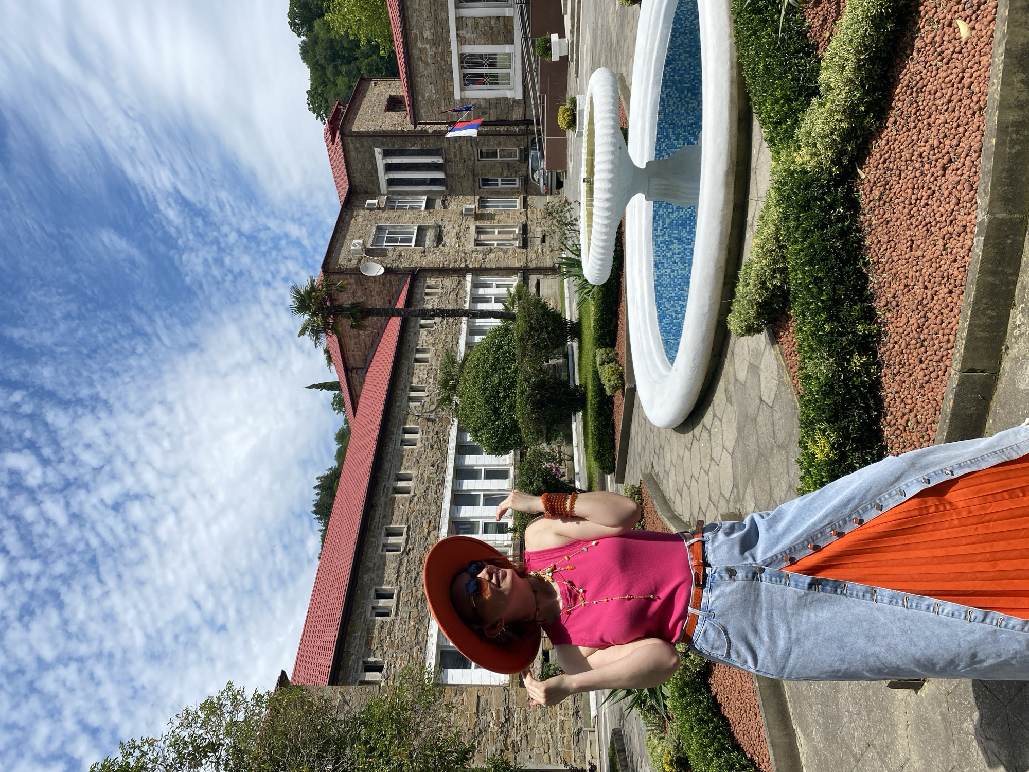The image depicts a woman standing in front of a building with a fountain in Sochi, Russia. The woman is wearing a pink top, a denim skirt with an orange pleated panel, a brown hat and sunglasses. She is wearing jewelry on her neck and wrists. The building behind her is a multi-story stone structure with a red tile roof. A fountain with blue tile is to the right of the woman. The sky above is blue with scattered clouds. Shrubs and trees add to the outdoor setting.