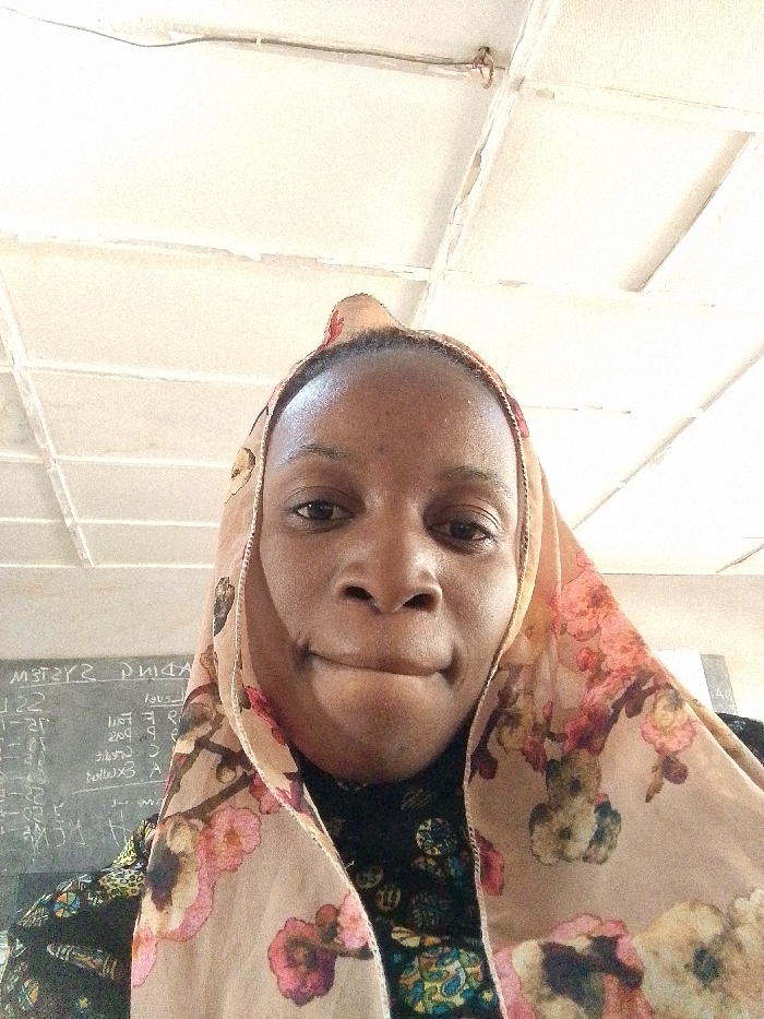 This image depicts a close-up, head-and-shoulders view of an adult female in an indoor setting within Barnawa, Nigeria. The woman, with a dark complexion, is centered in the frame, looking directly at the camera with a neutral expression and slightly pursed lips. She wears a light beige head covering, likely a hijab, adorned with a prominent floral pattern in shades of pink, orange, and green. A dark, patterned garment is visible underneath the head covering. The background on the left reveals a dark chalkboard with white, inverted writing, including partial words such as "SYSTEMS" and "ADING," and various numerals, suggesting a classroom environment. The upper background features a light-colored ceiling with a grid of square tiles, exhibiting some visible wear and a thin dark wire running across it.