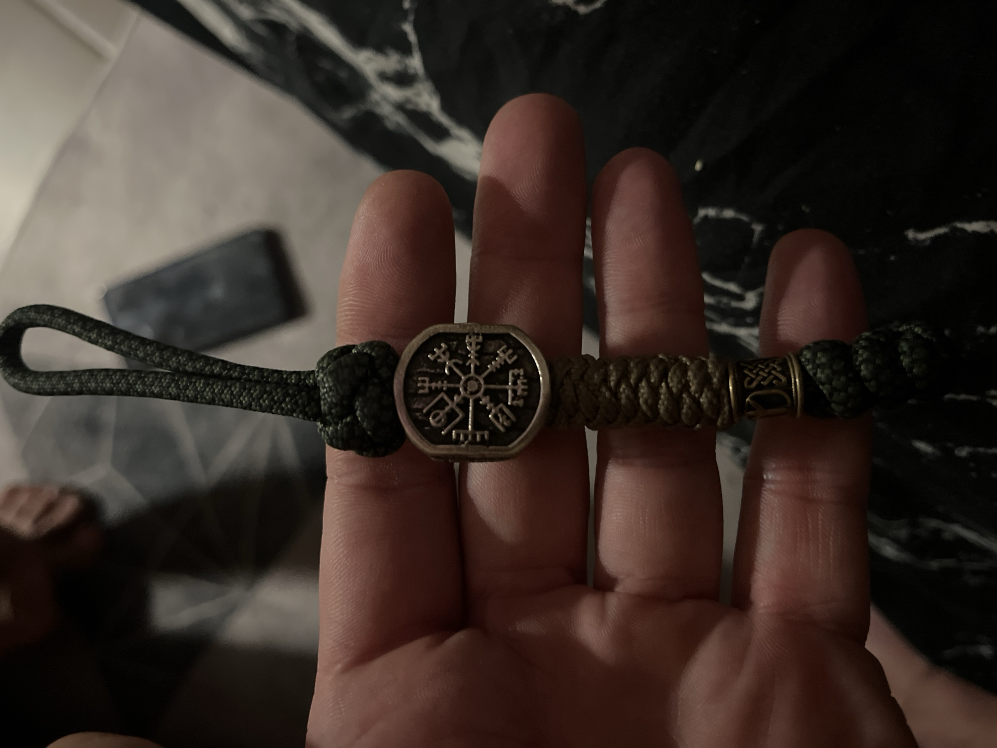 A close-up shot shows a hand holding a dark green paracord bracelet. The bracelet features a silver-colored charm in the center, depicting a compass-like symbol with runes. To the right of the charm, there's a section of braided tan paracord, followed by a small gold-colored decorative bead. The background consists of a dark, textured surface, possibly a bedspread or blanket, with a marble-like pattern. A blurred smartphone is visible in the upper left corner. The image is taken from a high-angle perspective. There is no discernible city or specific location information within the image itself, including Балаклавский округ, Ukraine.