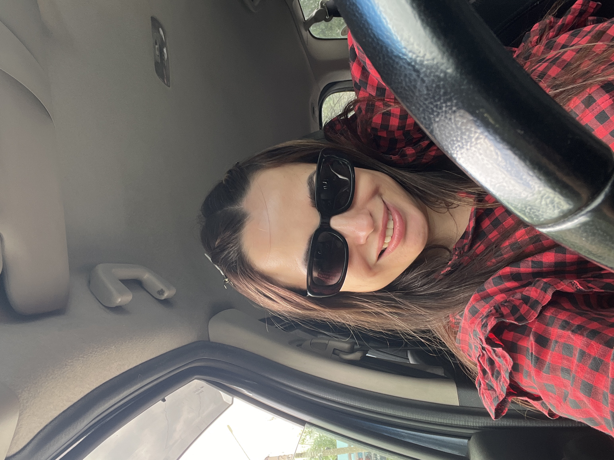 A young woman wearing sunglasses and a red and black checkered shirt is seated in the driver's seat of a car. She is smiling and looking at the camera. The steering wheel is visible in the foreground. Outside the car window, glimpses of buildings and greenery suggest a city environment, but the specific location is unknown.