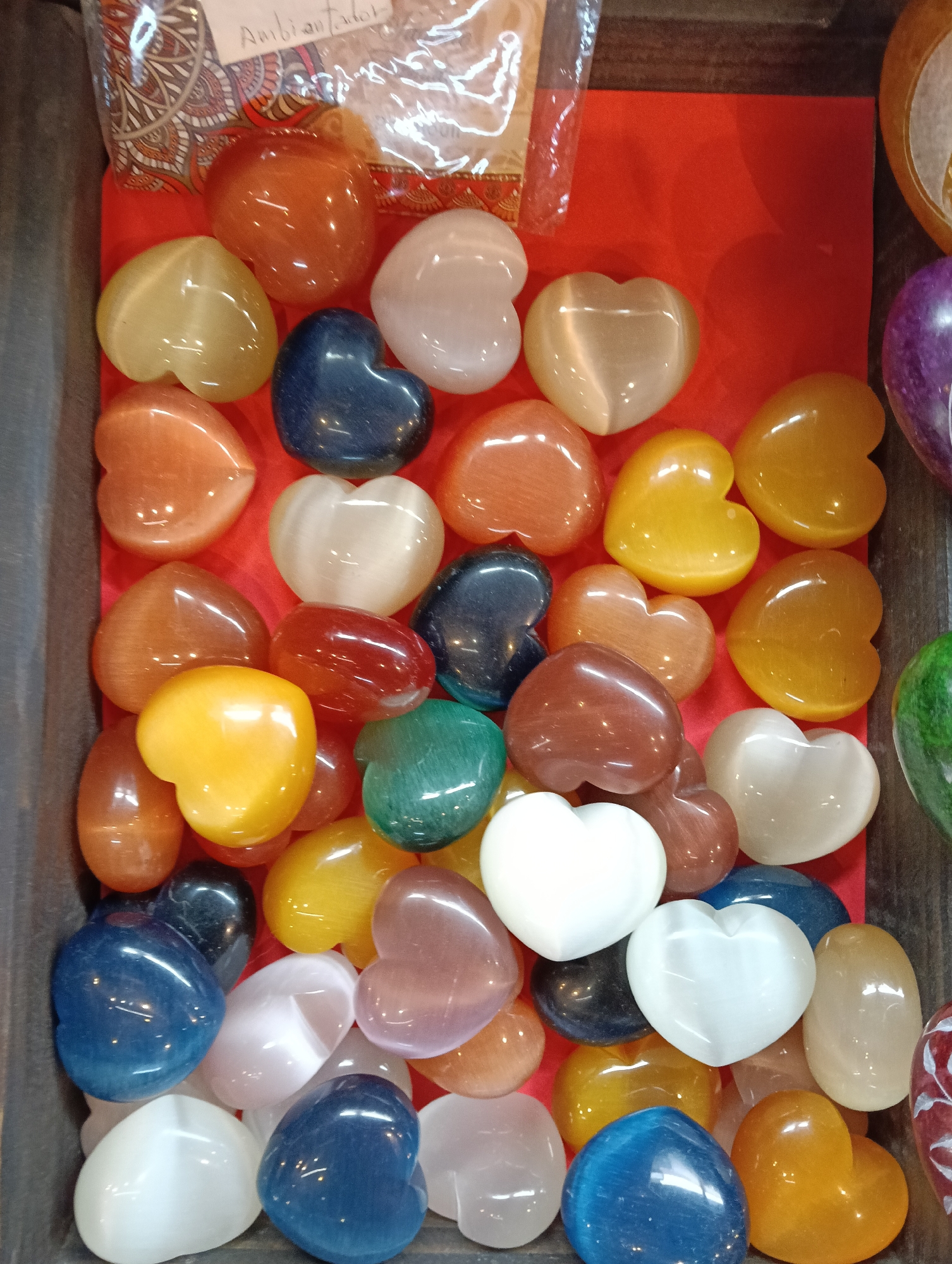 The image displays a collection of numerous polished, heart-shaped stones or crystals. These items are presented in a wide variety of colors, including vibrant shades of orange, yellow, dark blue, light blue, white, reddish-brown, and green, with some appearing translucent or exhibiting a cat's eye effect. A hint of purple can be seen on the right side.
The hearts are densely arranged on a bright red surface, which acts as a lining within a container, likely a wooden box or display tray. The smooth, shiny surfaces of the stones reflect light, creating bright highlights.
In the upper left corner, part of a plastic bag is visible, containing other items, possibly similar products. A piece of paper inside this bag clearly shows the Spanish word "Ambientador," meaning "air freshener." This text suggests that the bag contains air freshener products or items related to aromatherapy, which might be sold alongside the heart-shaped stones.
The scene depicts a display, indicating a retail environment such as a gift shop, market stall, or souvenir store, where these decorative or spiritual items are presented for sale. No people are visible, and there are no discernible details regarding the time of day or weather.