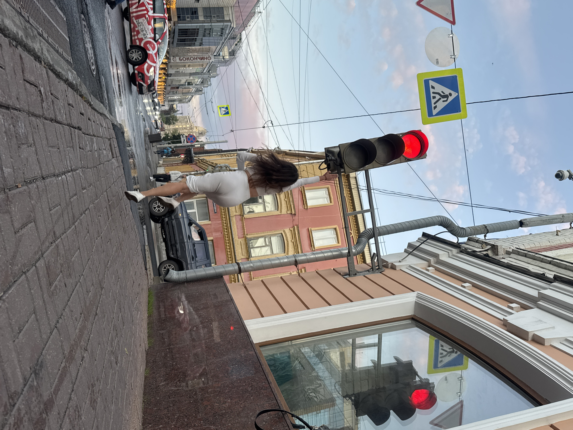 The image depicts a street scene in Nizhny Novgorod, Russia. A woman in white athletic wear is captured mid-jump or stride, her back to the camera, reaching upwards towards a traffic light that is illuminated red. She is positioned on a brick sidewalk next to a building with a pink facade and arched windows. Across the street, a red vehicle with white lettering is driving on a wet road, indicating recent rain. A dark SUV is parked further down the street. Overhead, traffic signals and wires are visible against a cloudy sky with hints of pink and orange from sunset. Road signs, including a pedestrian crossing sign, are also present. The urban environment suggests a commercial or residential street within the city.