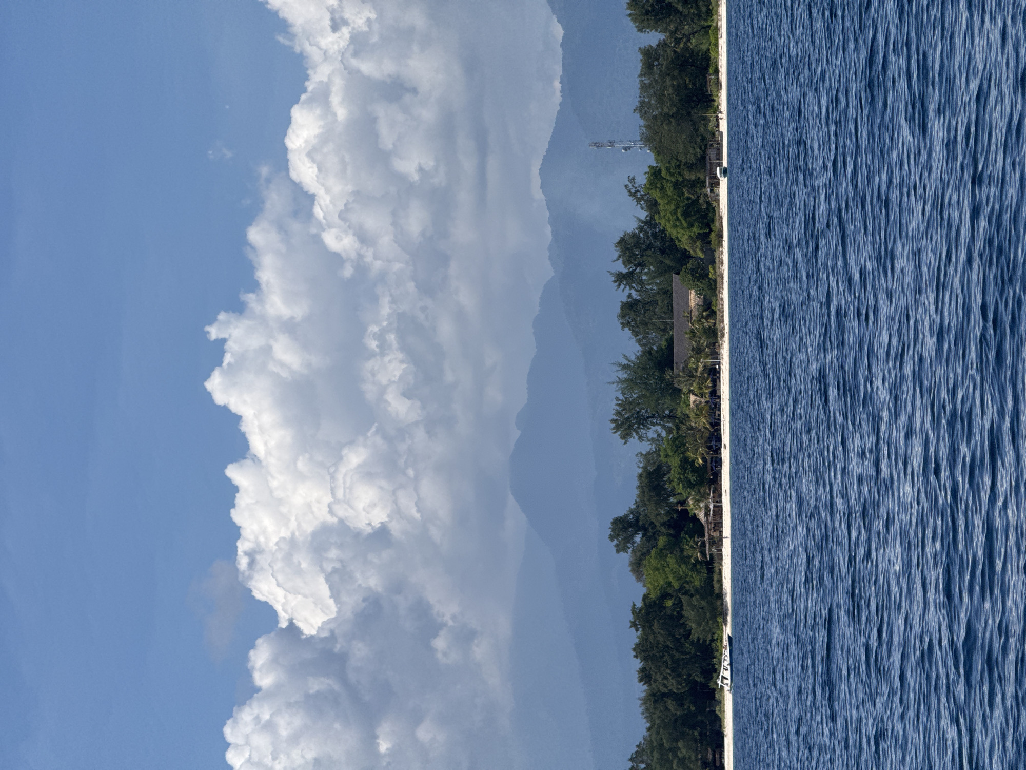 A landscape oriented image depicts a body of water on the right, with a shoreline lined with trees and buildings on the left-center. In the background behind the trees are hazy mountains. To the left of the mountains is a vast, cloudy sky. The image is rotated 90 degrees counterclockwise. The location is Unknown, Indonesia.