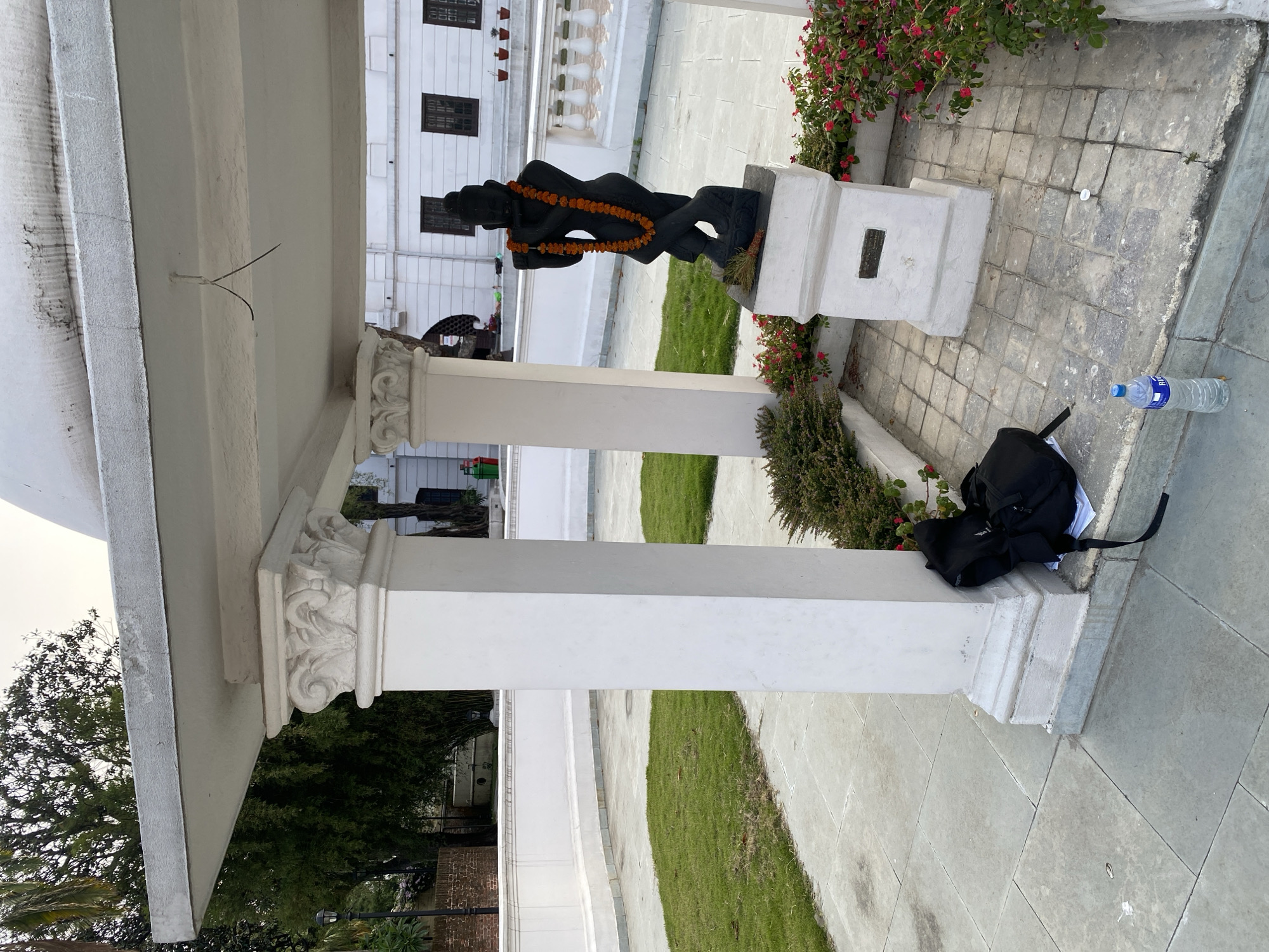 The image depicts a tranquil outdoor scene in Kathmandu Metropolitan City, Nepal, likely within a historical or cultural complex given the architecture. In the foreground, a black statue of a figure holding a flute stands on a pedestal, adorned with an orange marigold garland. Adjacent to the statue, a black backpack rests on the paved ground, near a clear plastic water bottle. The statue is situated within a decorative garden area with blooming pink flowers. Behind the statue and backpack, a white, classical-style pavilion with ornate columns provides shade. Beyond the pavilion, a white building with dark-framed windows is visible, suggesting a larger structure on the premises. The ground is paved with large stone tiles and interspersed with patches of lush green grass. Trees are visible in the background. The overall atmosphere is serene and hints at a place of cultural significance and public access.