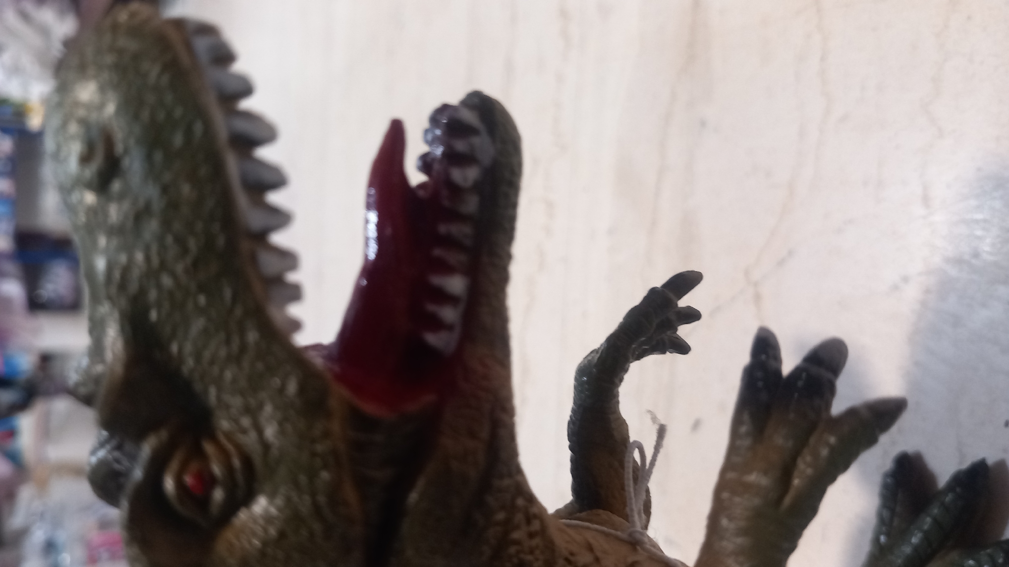 A close-up, blurred, eye-level shot shows the head and front claws of a toy Tyrannosaurus Rex. The dinosaur is dark green with red eyes and a gaping mouth revealing rows of sharp, white teeth and a red tongue. One of its clawed forearms is extended. The background is out of focus and appears to be a tiled surface, possibly a tabletop, with some shelves and objects visible in the distance. There is no indication of the city of Kos, Greece, in this image.