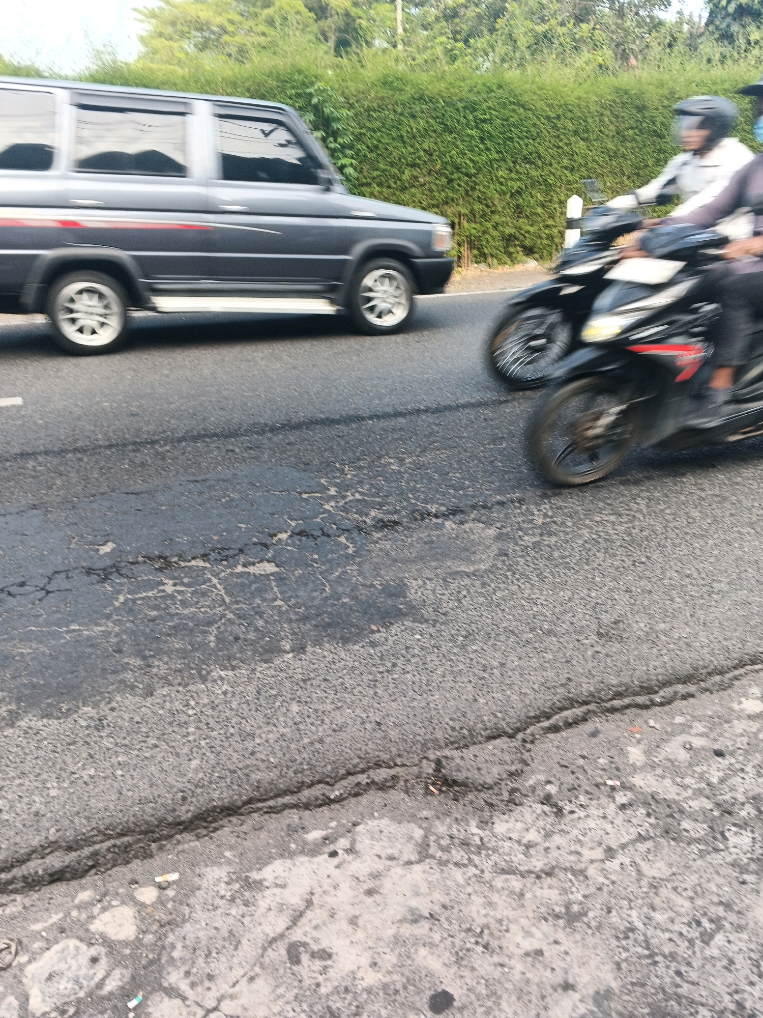 In Ungaran, Indonesia, a dark gray SUV is traveling on an asphalt road, partially obscured by motion blur. To its right, two individuals on a black motorcycle are also moving in the same direction. The motorcycle rider in front wears a white jacket and a dark helmet, while the passenger behind them is dressed in dark clothing. The road surface exhibits signs of wear, with visible cracks and patches. A dense, green hedge forms the background, and a white post stands beside the road. The lighting suggests it is daytime.