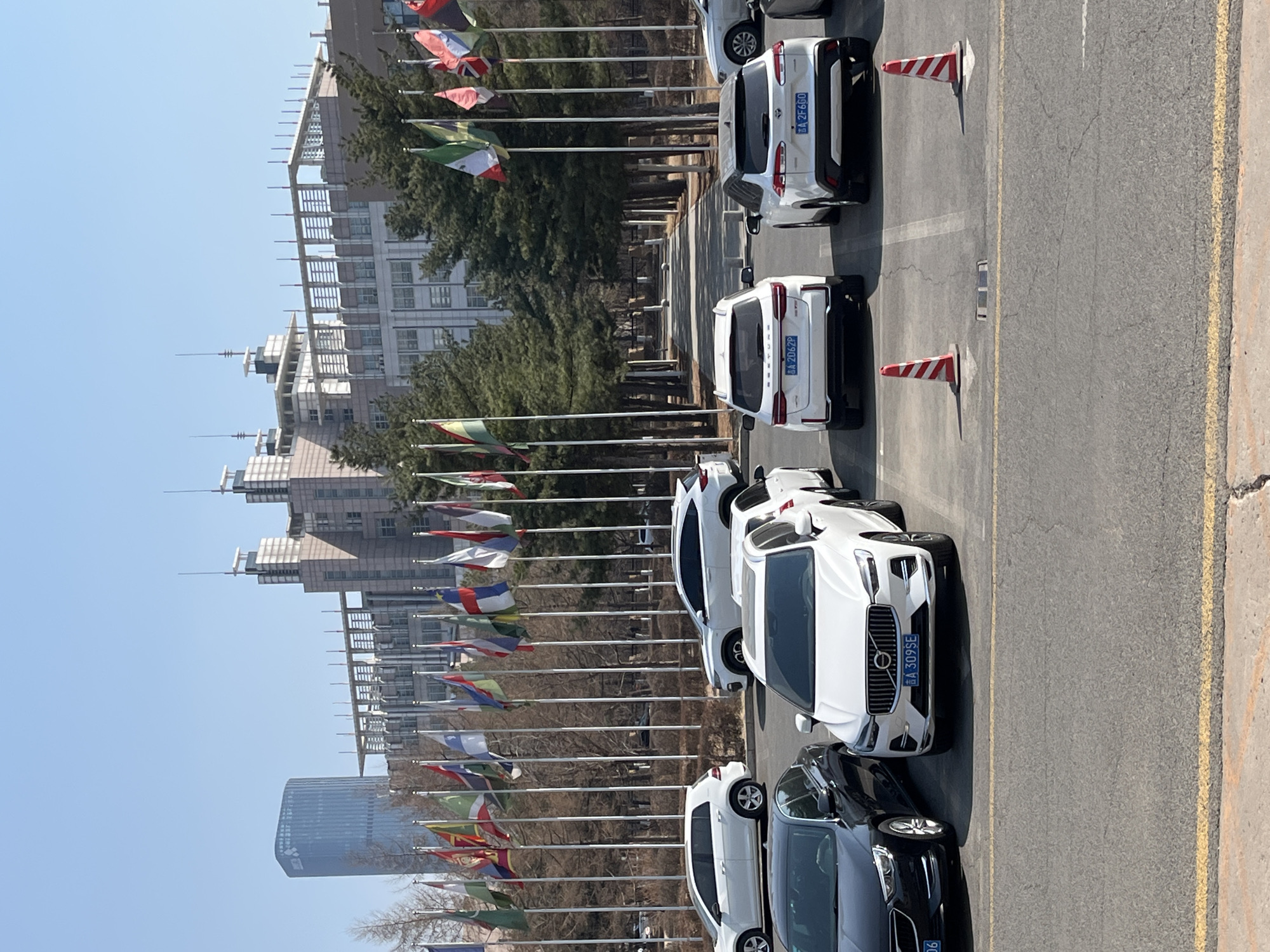 The image shows a street in Changchun City, China lined with international flags. On the right side of the image, cars are parked, interspersed with traffic cones. Buildings can be seen in the background, along with some trees. The sky is clear and blue.