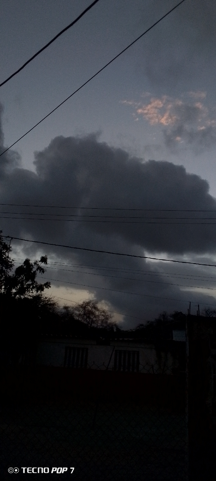 The image shows a dark, overcast sky with heavy clouds, some with a hint of pink light. Power lines are visible against the sky. Below the sky, there is a building with windows, obscured by shadows and foliage. The image was taken in Altagracia, Venezuela, but specific details about the location are not discernible. The scene is dark, and the foreground is mostly obscured. The camera used was a TECNO POP 7.