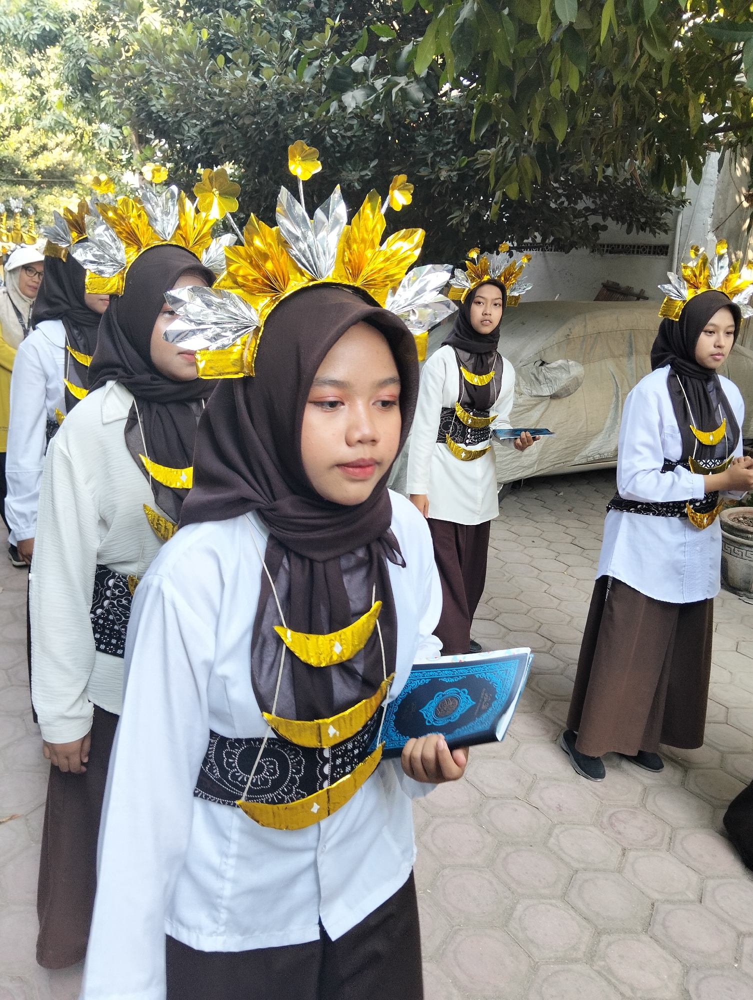 In Kranji, Indonesia, a group of young girls in hijabs and formal attire, likely part of a school event, are captured in a procession. The girls are adorned with elaborate, golden and silver headpieces that resemble fan-like headdresses, adding a festive and ceremonial air to the scene. They are wearing white shirts and brown skirts or trousers, with decorative bands around their waists. One girl in the foreground is holding an open book, possibly a religious text given the intricate blue cover with Arabic calligraphy, suggesting the event might be related to a religious or cultural celebration. The ground is paved with hexagonal tiles, and lush green foliage forms a natural backdrop, indicating an outdoor setting, possibly within the grounds of an educational institution or a community gathering space. The girls appear to be walking in an organized line, facing forward with a sense of purpose and engagement.