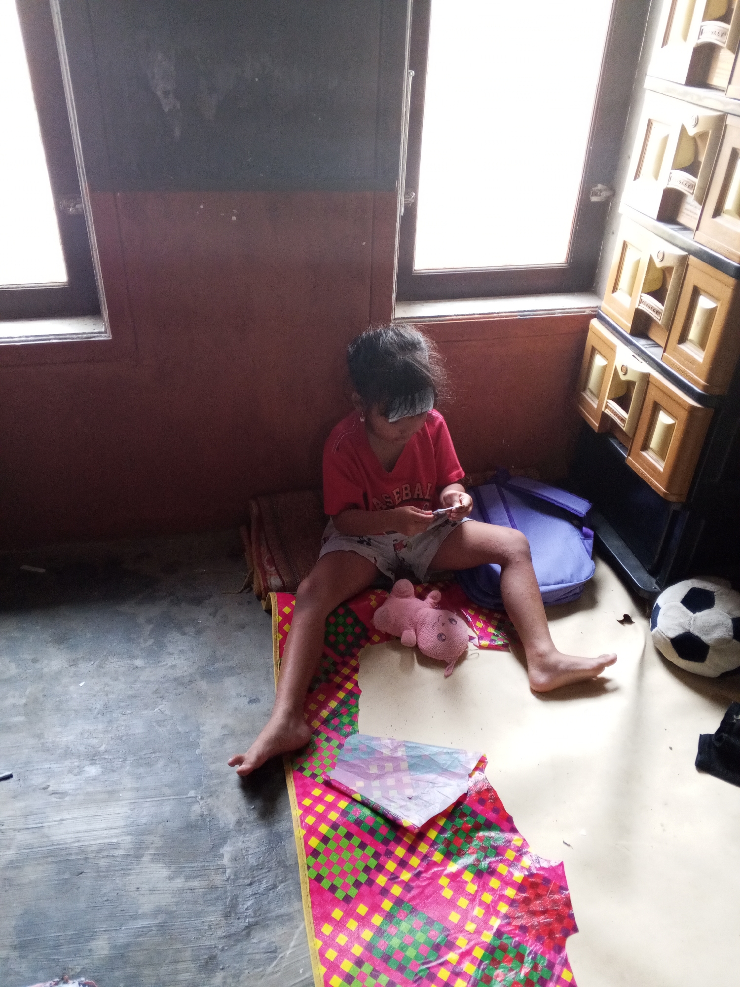 A young child, appearing to be of East Asian descent, sits on the floor in a room. The child is wearing a red t-shirt and patterned shorts, and is holding a small white object, possibly a phone, in their hands. To the child's left is a pink stuffed animal, and to the child's right is a soccer ball. The room appears to be dimly lit, with natural light entering from two windows on the left side of the image. A tall, multi-drawer plastic cabinet is visible to the right of the child. The floor is a mix of concrete and what appears to be a patterned mat. The scene is consistent with an indoor residential setting in Turen, Indonesia.