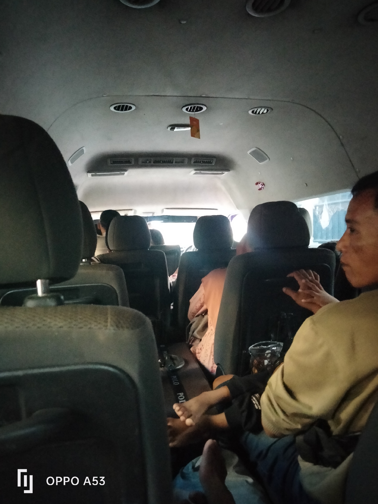 The image, captured within a van, shows several individuals seated. In the foreground, on the right, a man in a light brown jacket is visible in profile, his right hand resting on the back of a headrest. His face is turned to the left, suggesting he is looking at something or someone within the van. Partially obscured by him, and further back in the van, are other passengers. To the left of the frame, the back of a dark grey headrest is prominent. The van's interior features an overhead console with air vents and lights. A strap with the word "POLO" printed on it is visible near the floor. A glass with a dark liquid is placed on a surface between the seats. The overall scene suggests travel within a confined space. The location is identified as Panempan, Indonesia.