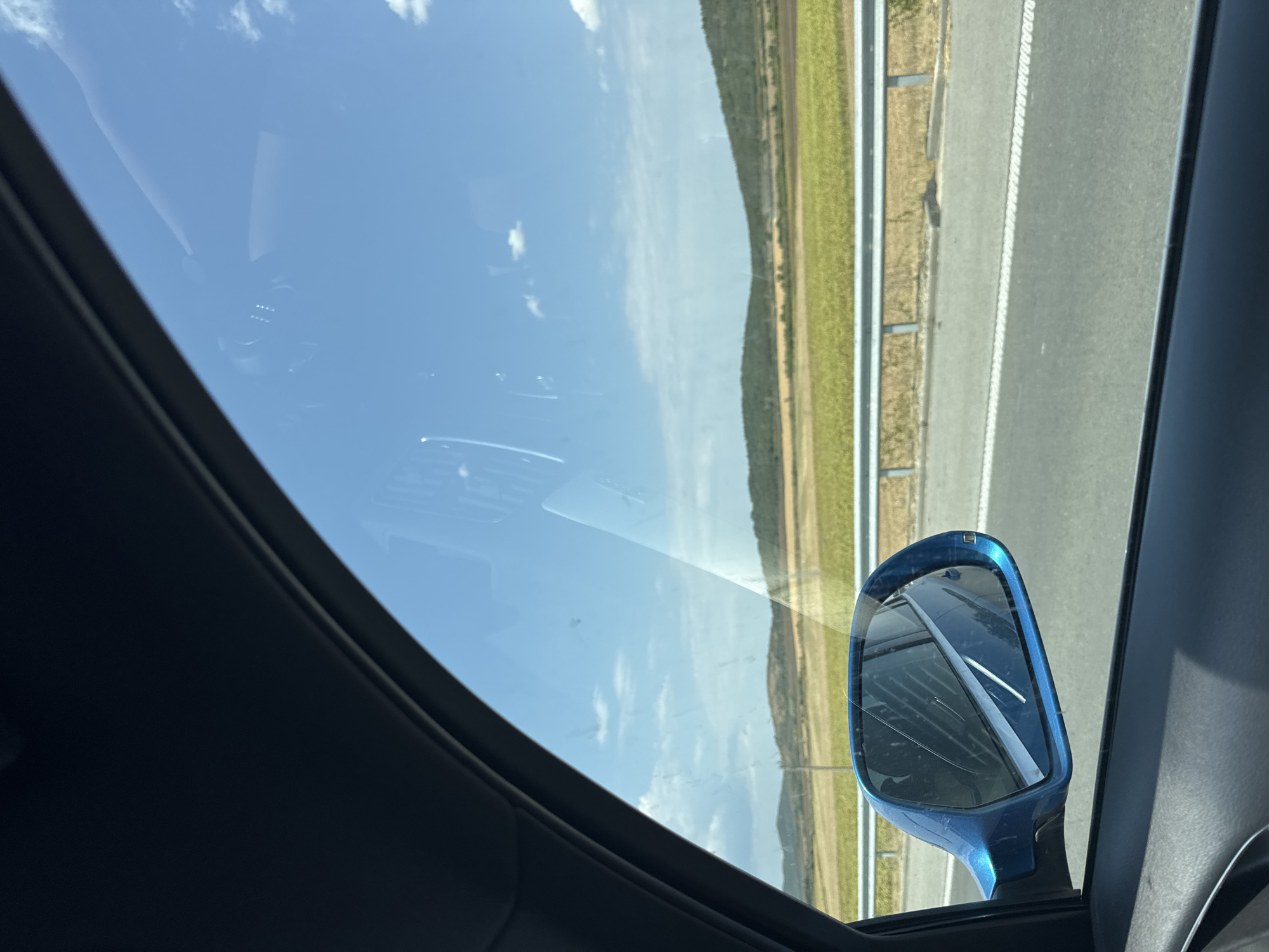 The image, captured from inside a vehicle, offers a view through a car window. The primary focus is the side mirror, which is a bright blue and reflects the car's interior and the exterior landscape. The exterior scene includes a highway with a metal guardrail and a vast expanse of agricultural fields and rolling hills under a clear blue sky with scattered white clouds. The scene appears to be located in the vicinity of Kaloyanovets, Bulgaria, given the agricultural landscape. There are no people visible in the image, and the action is static, representing a moment in time during a drive. The environment is characterized by rural scenery and a sunny day.