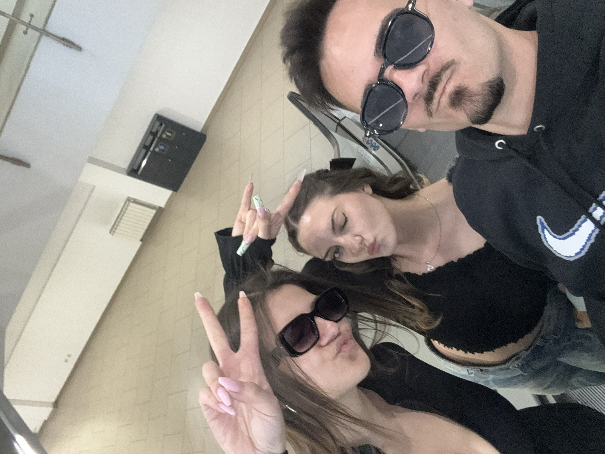 Three people are in the photograph, possibly taking a selfie while on an escalator. Two women are making peace signs with their hands. One woman is wearing sunglasses. The man is wearing sunglasses and a black hooded sweatshirt. The background features a tiled floor, and a black control panel mounted on the wall. The location is likely in Sofia, Bulgaria, but this is not definitively evident in the photograph.